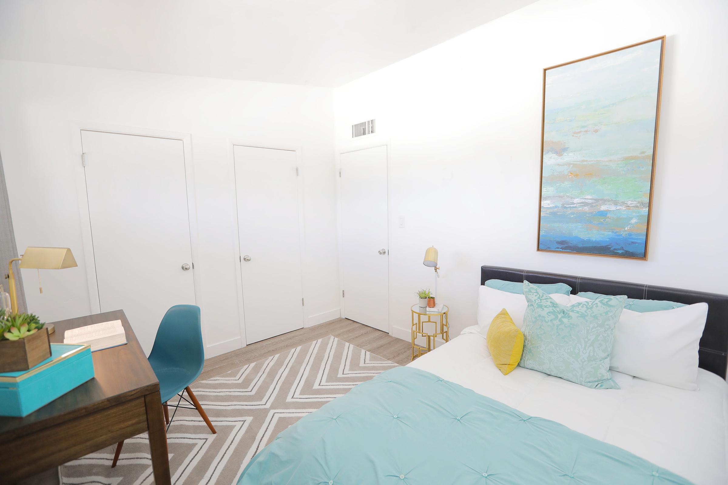 A modern bedroom featuring a bed with light blue and white bedding, a decorative yellow pillow, and a colorful abstract painting above the headboard. The room includes a wooden desk with a lamp, an aqua chair, and a small plant. Two white closet doors are visible in the background, along with a neutral patterned rug.