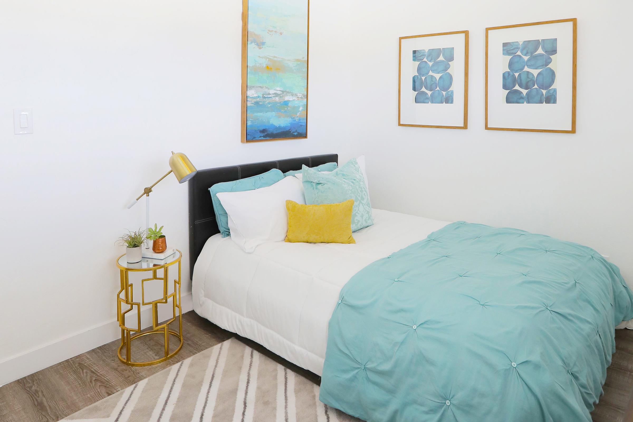 A modern bedroom featuring a black bed with a white comforter and turquoise accent pillows. A small round side table with a gold lamp is next to the bed, adorned with a plant. Two framed artworks with blue and abstract designs hang on the wall, complementing the overall decor.