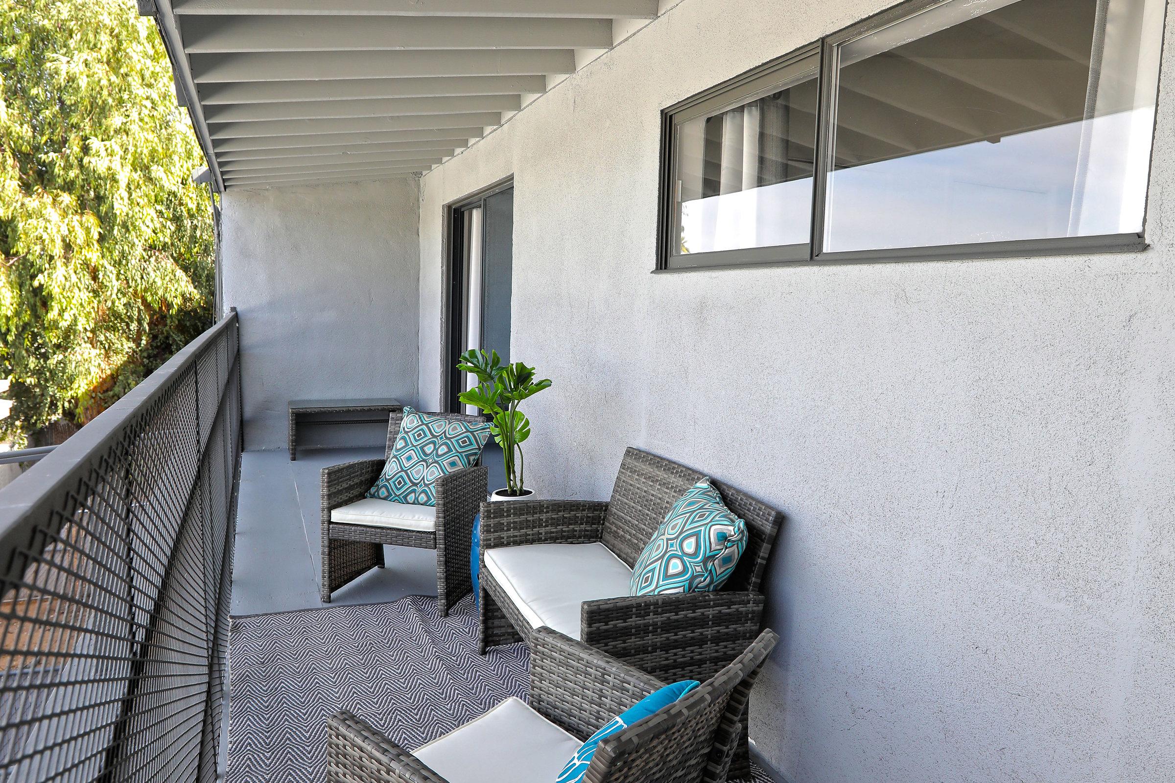 A balcony featuring two wicker chairs with decorative cushions, a small potted plant on a table, and a patterned rug. Light from a window and greenery outside enhance the cozy atmosphere.