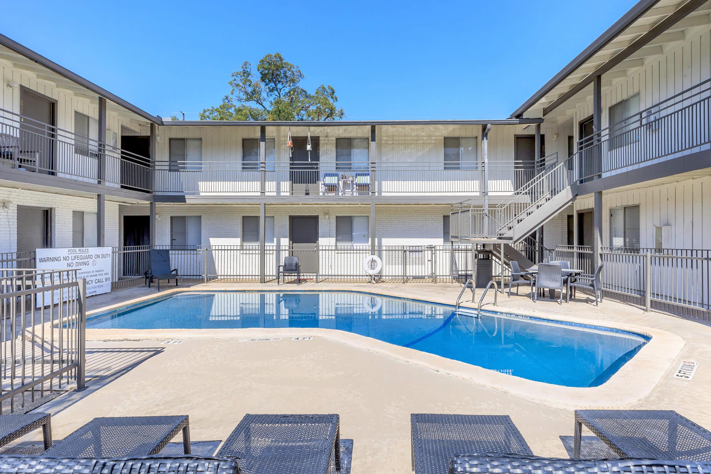 A view of a multi-story apartment complex featuring an outdoor swimming pool in the center. The pool area includes lounge chairs and a surrounding railing. The building has several balconies visible on the upper level, with trees in the background under a clear blue sky.