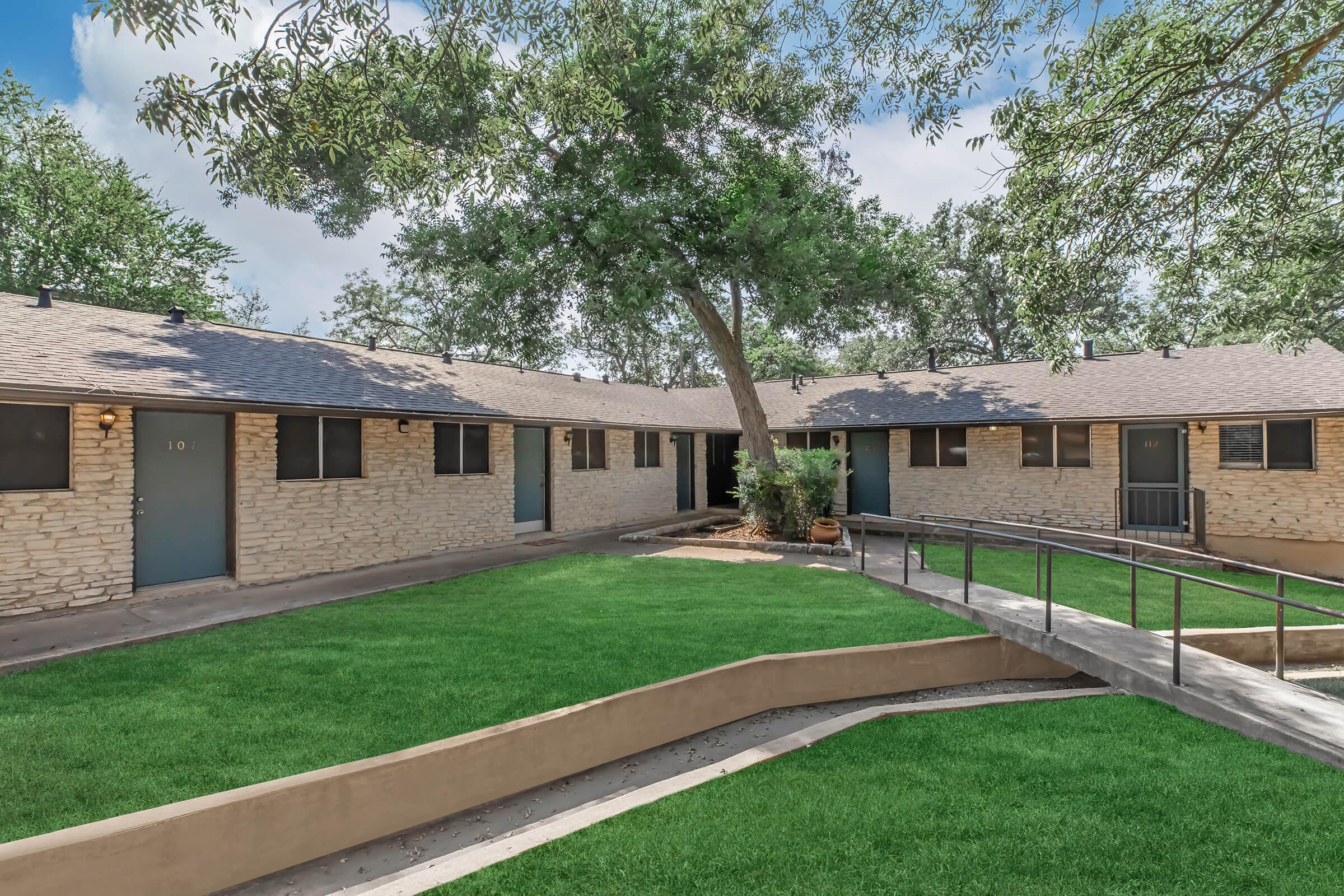 A view of a landscaped area featuring several single-story buildings made of light-colored stone. Each unit has a door, and the scene is surrounded by greenery and trees. A gently sloping pathway leads to the entrance, highlighting a well-maintained lawn in a serene environment.