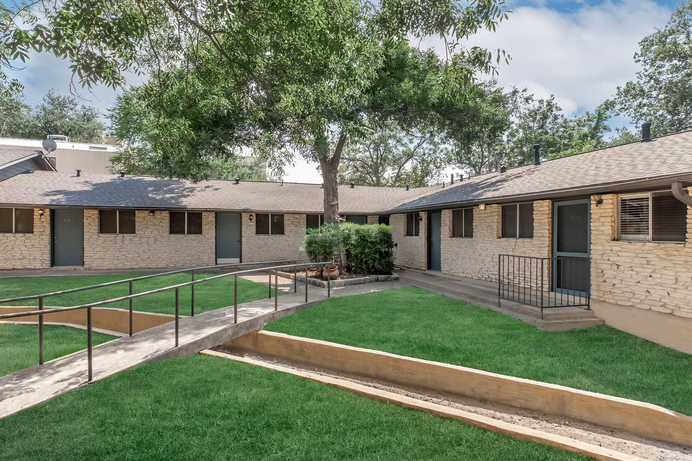 A well-maintained, two-story building featuring light-colored stone exteriors and black doors. The surrounding area has lush green lawns and a pathway with gentle slopes. Trees provide shade, enhancing the inviting atmosphere of this residential complex.