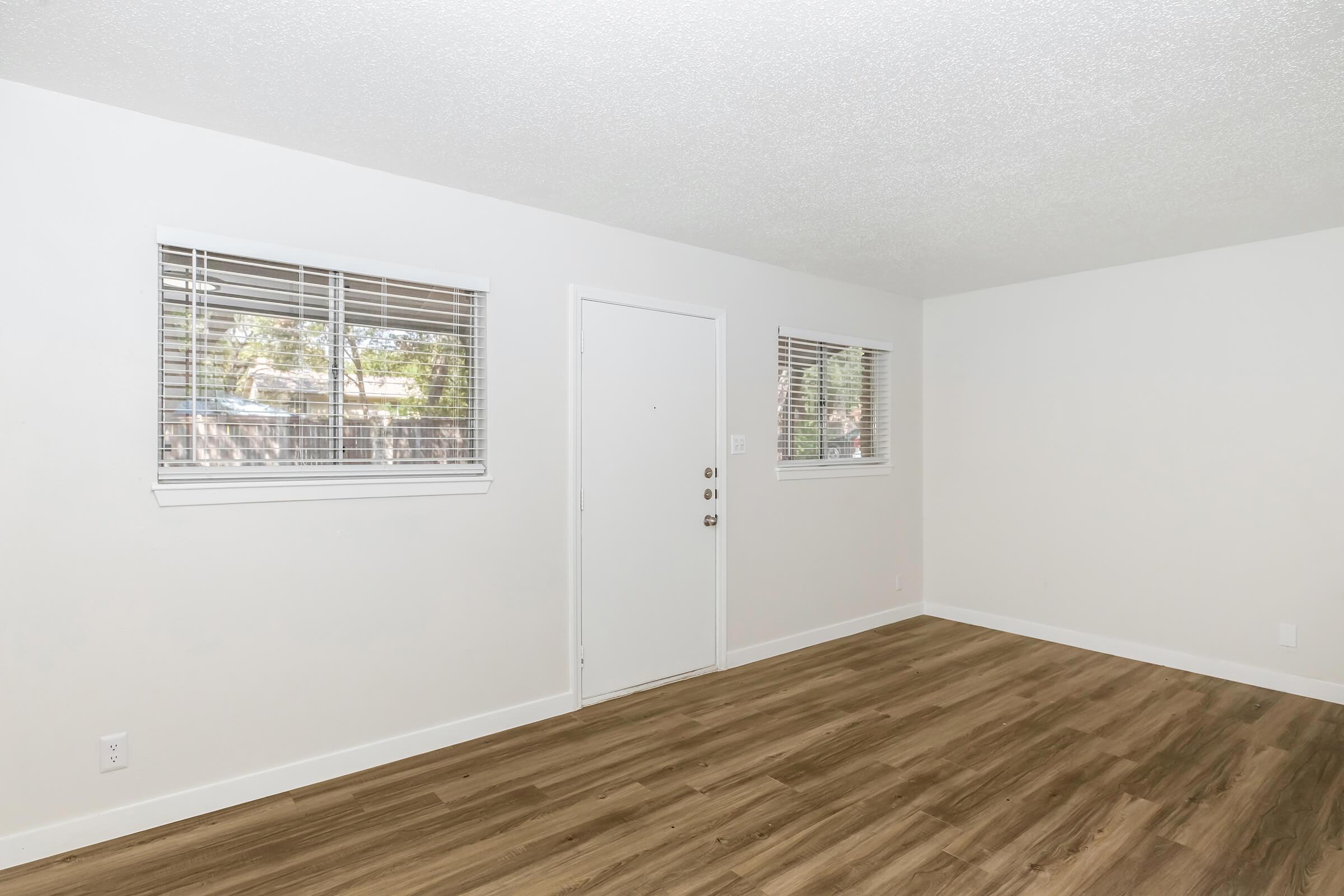 An empty room with light-colored walls and brown wooden flooring. Two windows with blinds allow natural light, and a white door is centered on the wall. The space has a clean and minimalist aesthetic, suitable for various uses or furnishings.