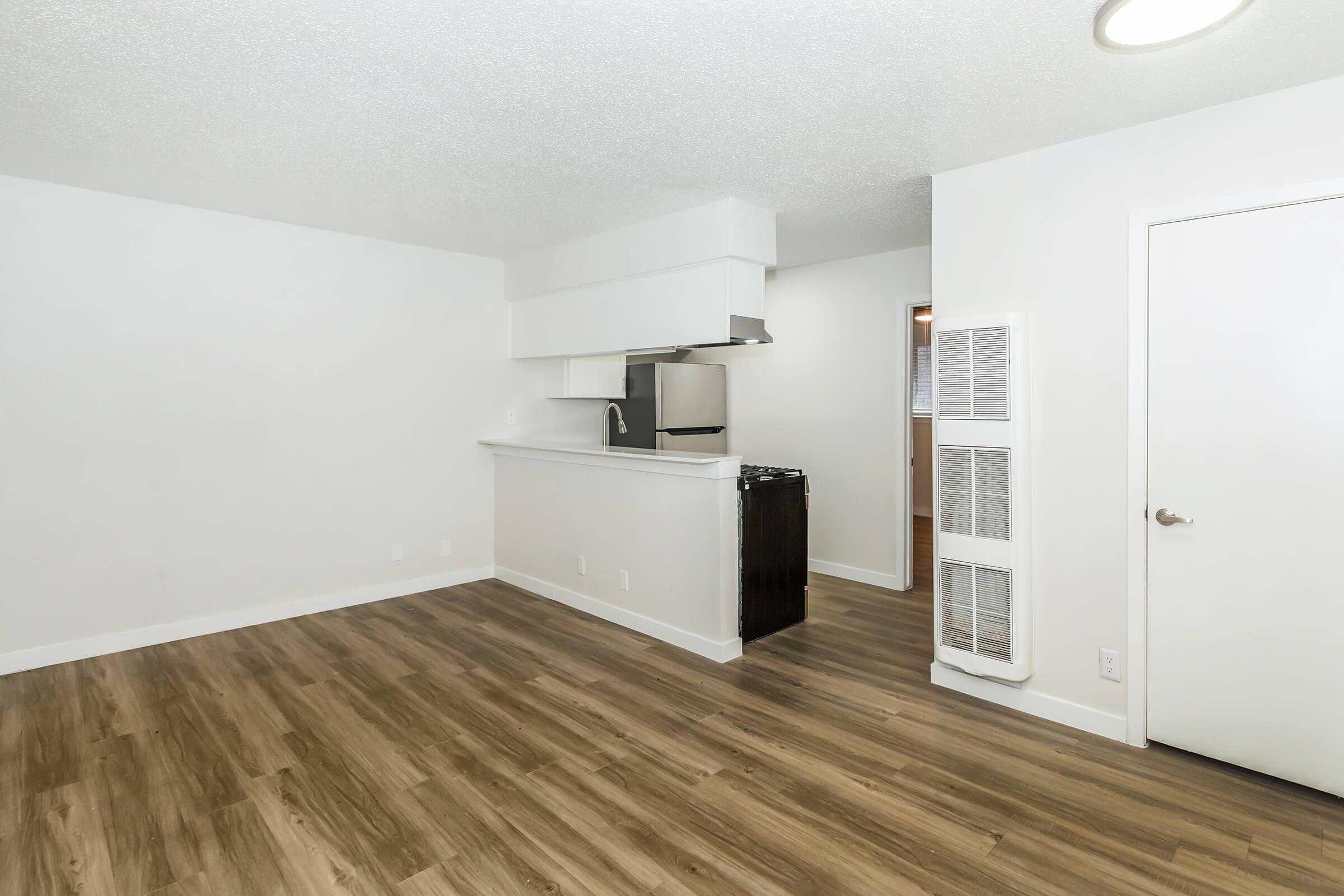 A modern, empty apartment interior featuring light-colored walls and a wood-like laminate floor. The kitchen area has a compact setup with an overhead hood, stainless steel appliances, and a small breakfast bar. The room is well-lit, creating an open and spacious atmosphere.