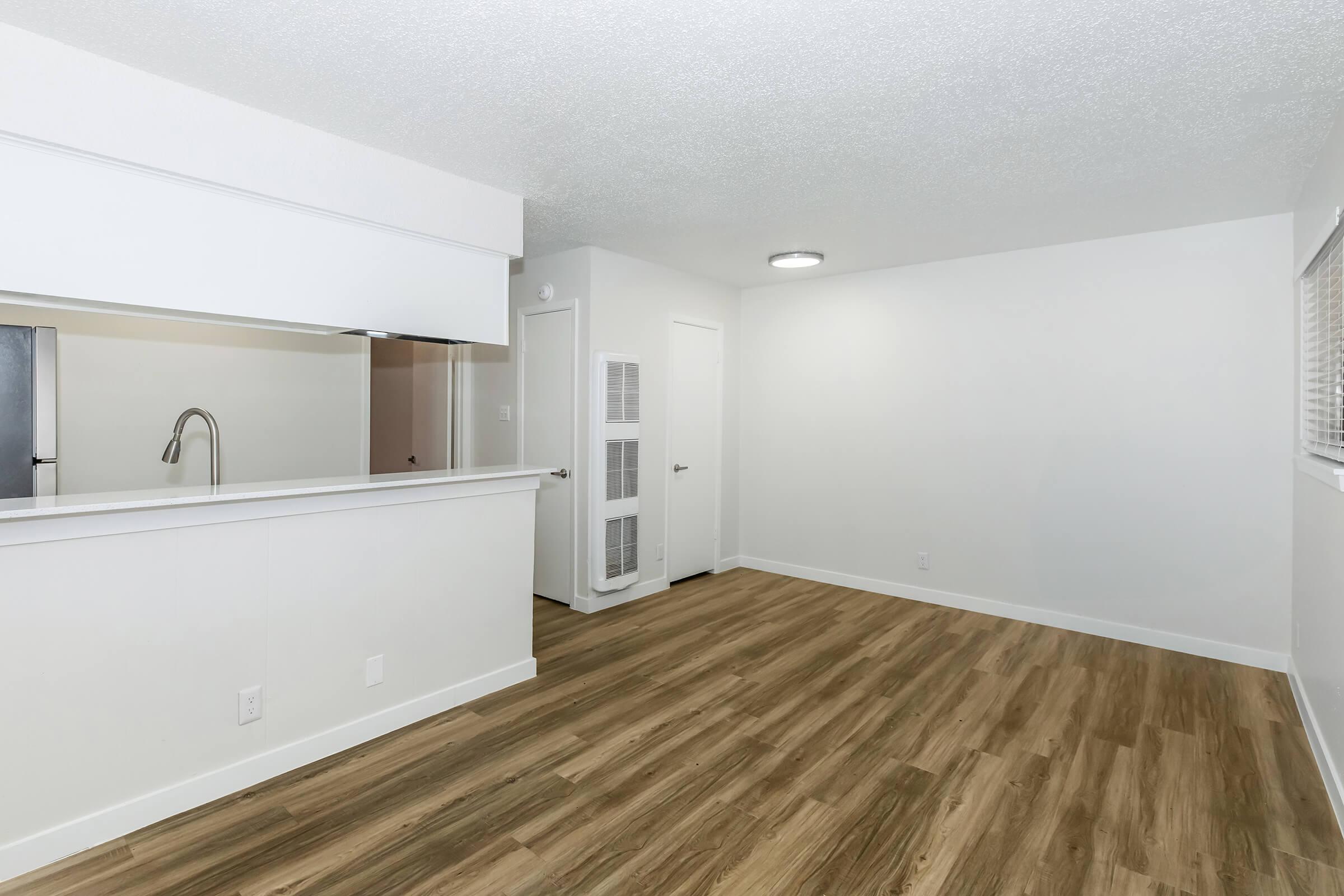 A spacious, modern living area with light wood flooring. The room features white walls and a large window letting in natural light. An open kitchen with a bar-style counter is visible, along with a doorway leading to another room and a closet with mirrored doors. The overall ambiance is clean and bright.