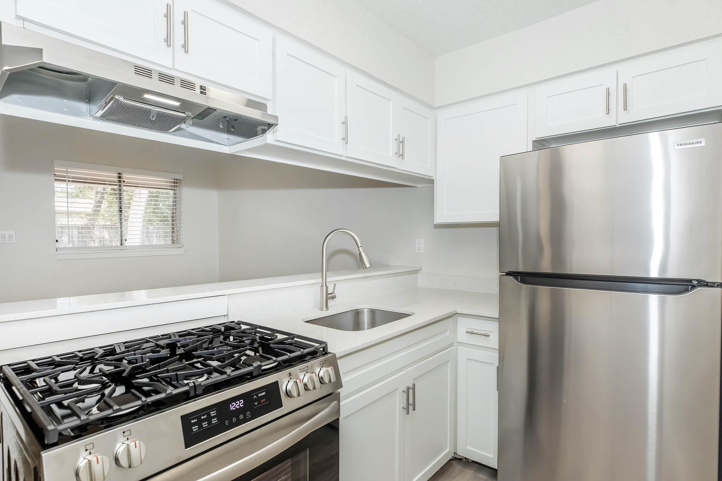 Modern kitchen featuring stainless steel appliances, including a gas range and refrigerator. White cabinetry and a small sink complement the light-colored countertops. A window offers natural light. The overall design is clean and contemporary, suitable for efficient cooking and dining.