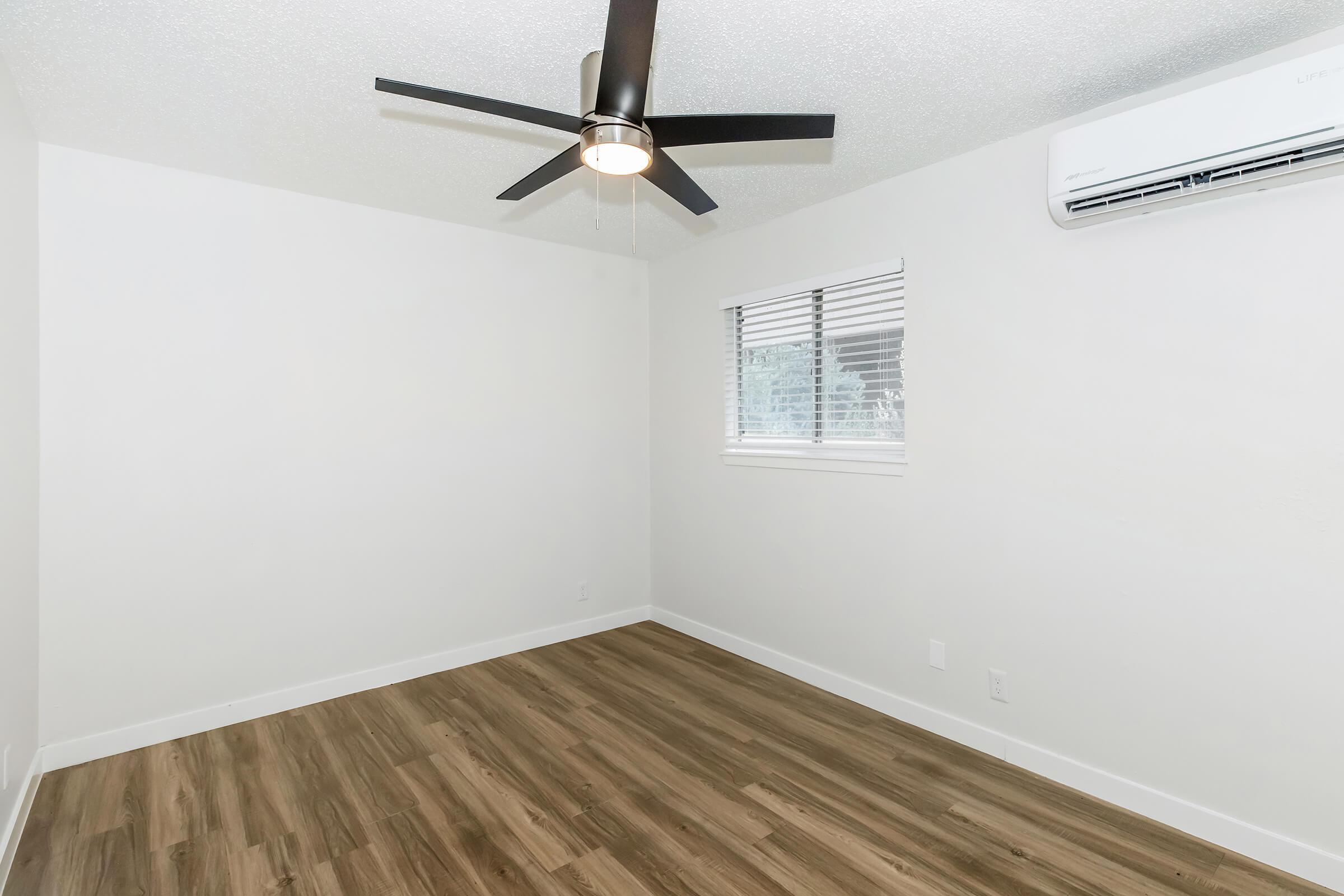 Empty, well-lit room featuring a ceiling fan and a window with blinds. The hardwood floor complements the neutral-colored walls, and an air conditioning unit is mounted on the wall. The overall aesthetic is modern and minimalistic.
