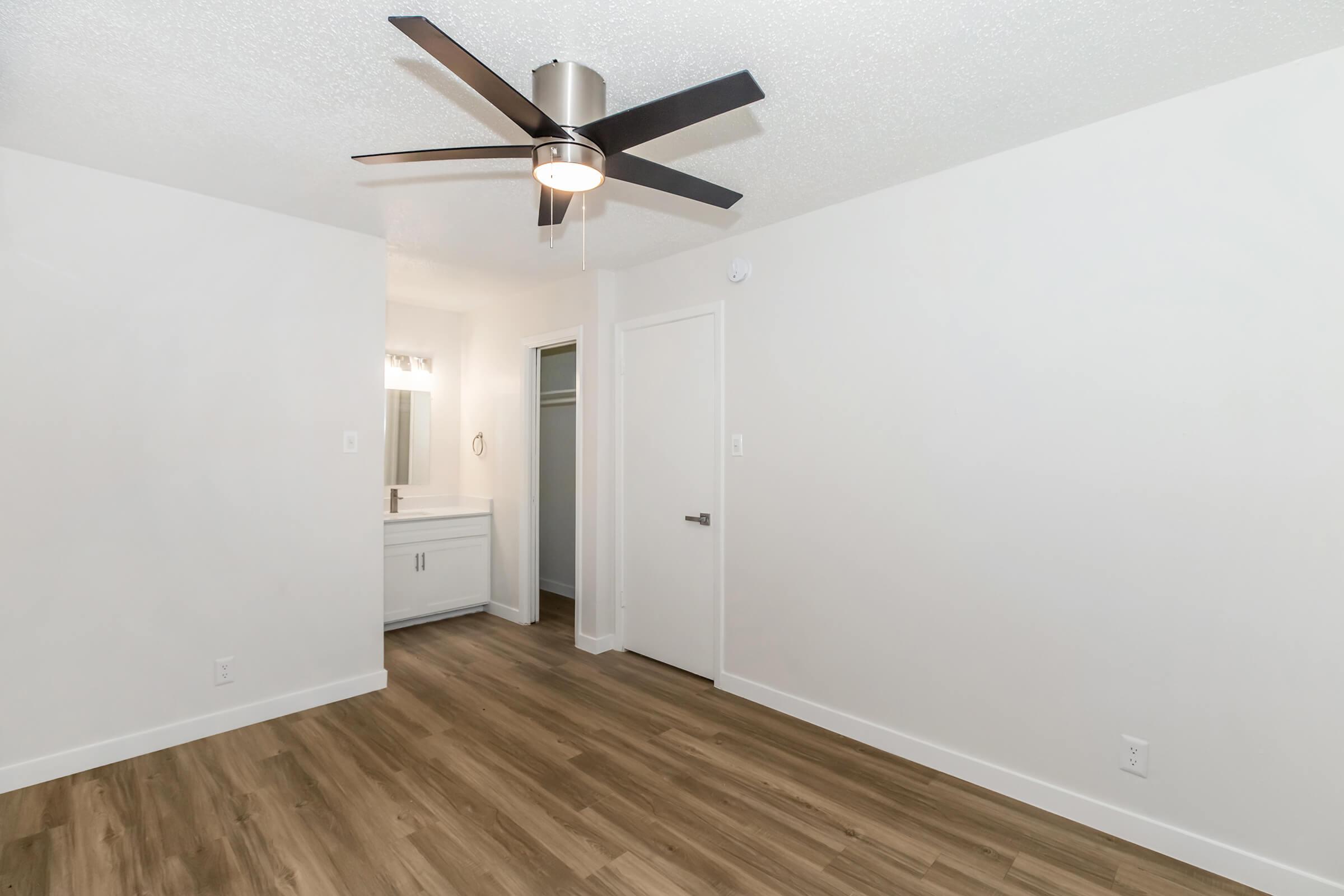 A well-lit interior of a modern room featuring light-colored walls, a ceiling fan, and hardwood flooring. There is a doorway leading to a bathroom area with a sink and mirror. The room has a minimalist design, enhancing the spacious feel.