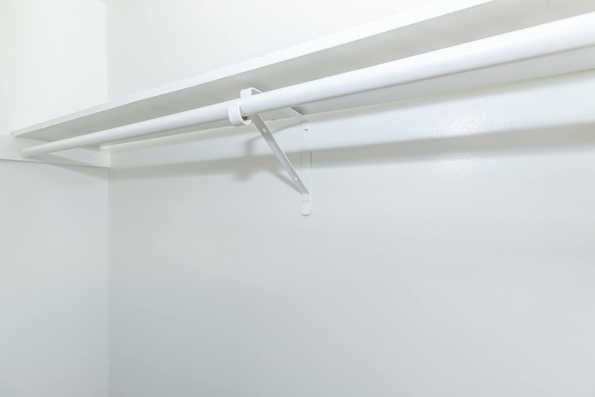 A partially empty closet with a white wall and a simple white clothes rod. There is one hook hanging down from the rod, indicating a minimalistic design. The overall space appears clean and uncluttered.