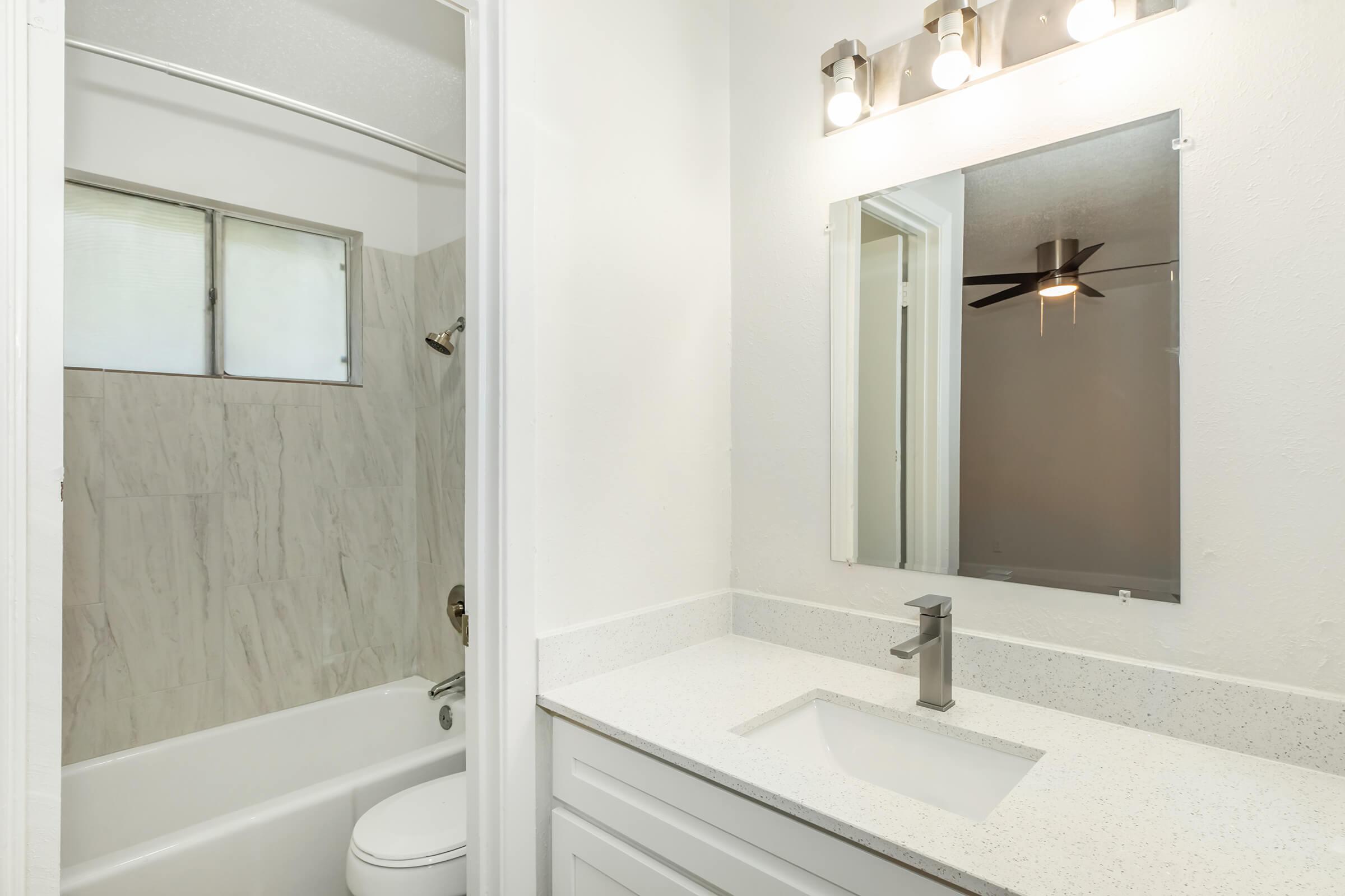 Bright and modern bathroom featuring a white marble-like tile shower, a clean bathtub, a sleek countertop with a silver faucet, a well-lit mirror, and a ceiling fan. The walls are painted in neutral tones, enhancing the spacious feel of the area. Natural light comes through a window above the tub.