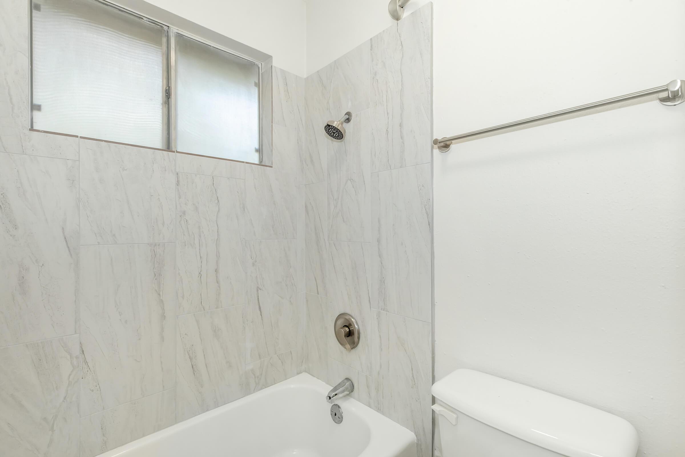 A modern bathroom featuring a white bathtub with a showerhead above, a sleek toilet, and light-colored marble wall tiles. A small window is visible above the bathtub, allowing natural light to enter the space. The overall design is minimalist and clean.