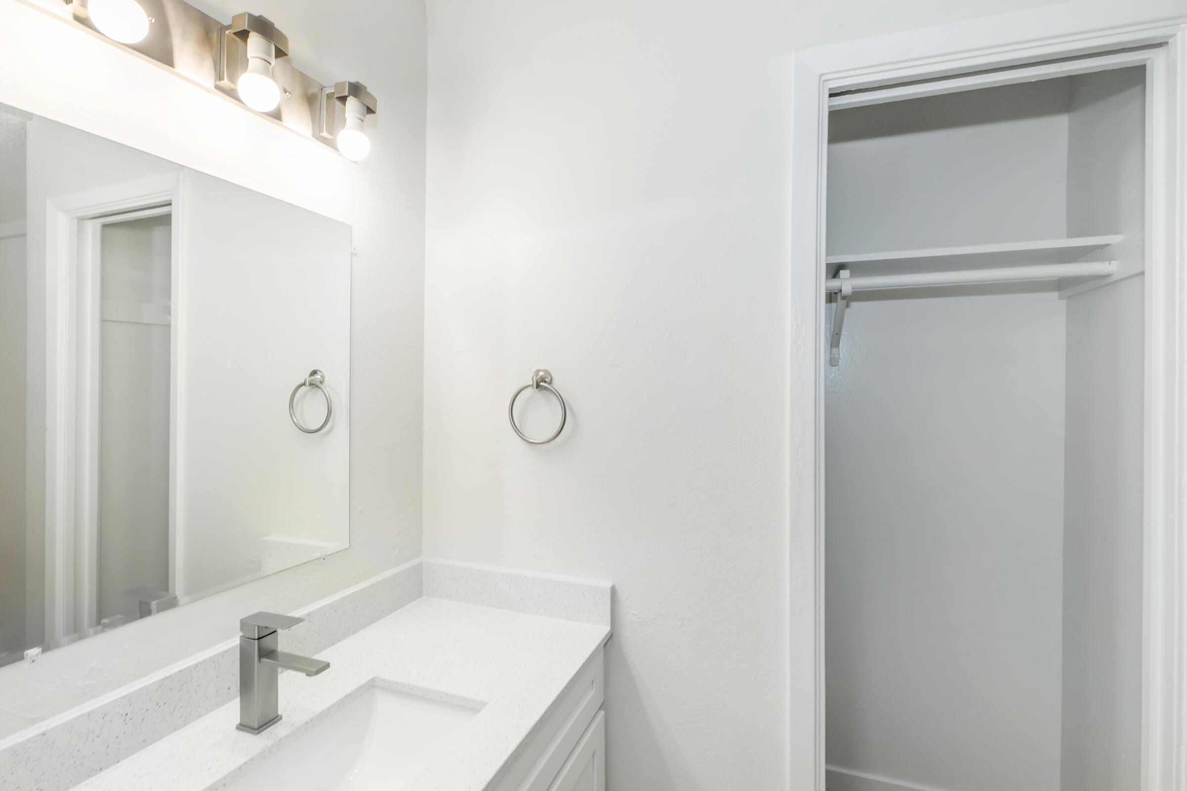 A modern bathroom featuring a white vanity with a rectangular sink, sleek faucet, and a round towel ring mounted on the wall. Above the sink, there is a large mirror reflecting the bright, well-lit space. To the right, an empty closet with a shelf and hanging bar is visible. The walls are painted white.
