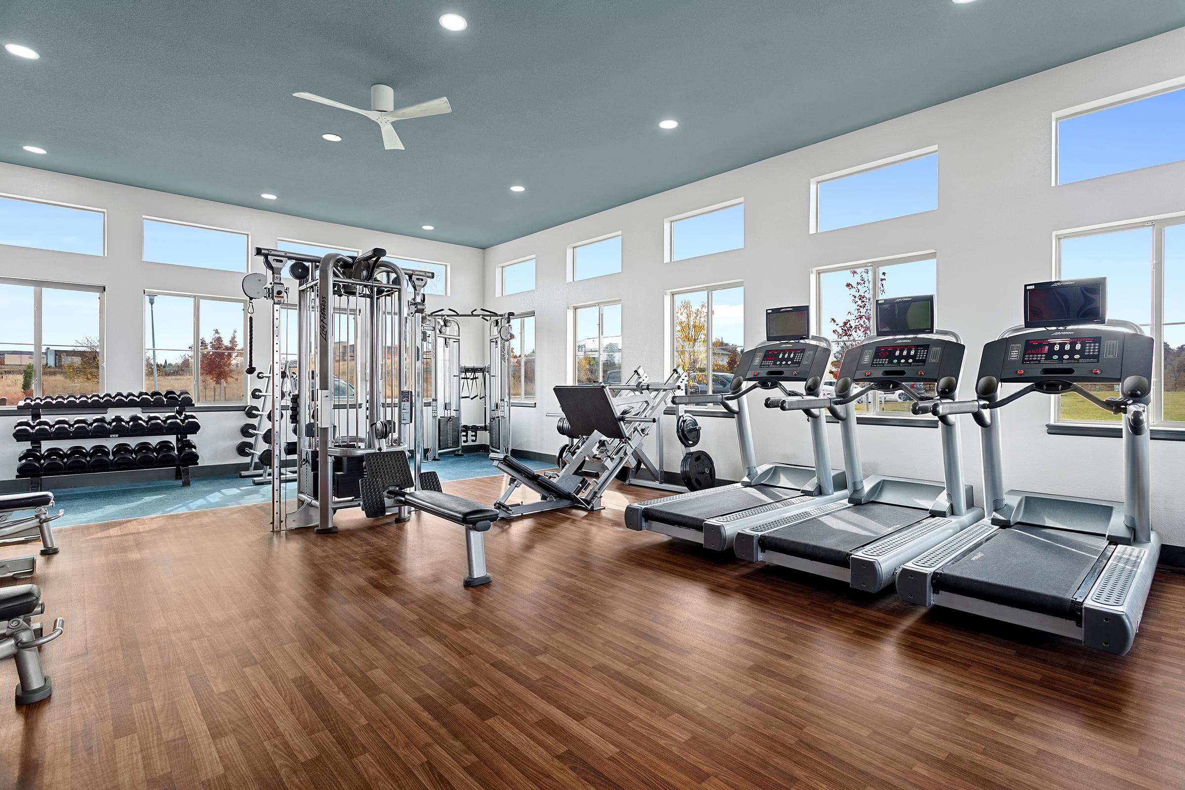 A spacious, well-lit gym featuring modern fitness equipment. There are treadmill machines, a weight bench, and multi-functional weight machines. Large windows provide natural light, and the floor is made of hardwood. The environment is clean and organized, promoting a welcoming atmosphere for exercise.