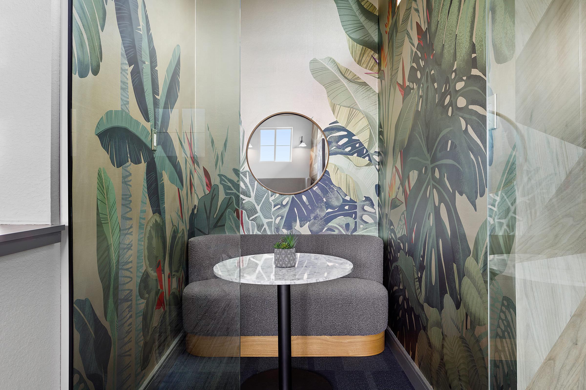 A modern, cozy meeting space featuring a curved gray sofa and a round marble-top table. The walls are adorned with vibrant tropical foliage murals, and a circular mirror reflects light. Natural light enters through a window, creating a welcoming atmosphere. A small green plant adds a touch of nature.