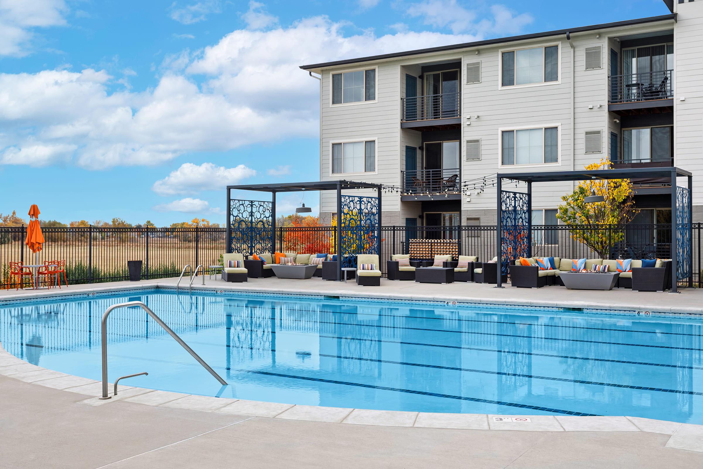 A clean, blue swimming pool surrounded by lounge seating and decorative cabanas. The scene includes a modern apartment building in the background, with orange umbrellas and autumn-colored trees nearby, all set against a clear sky. The area is perfect for relaxation and leisure.