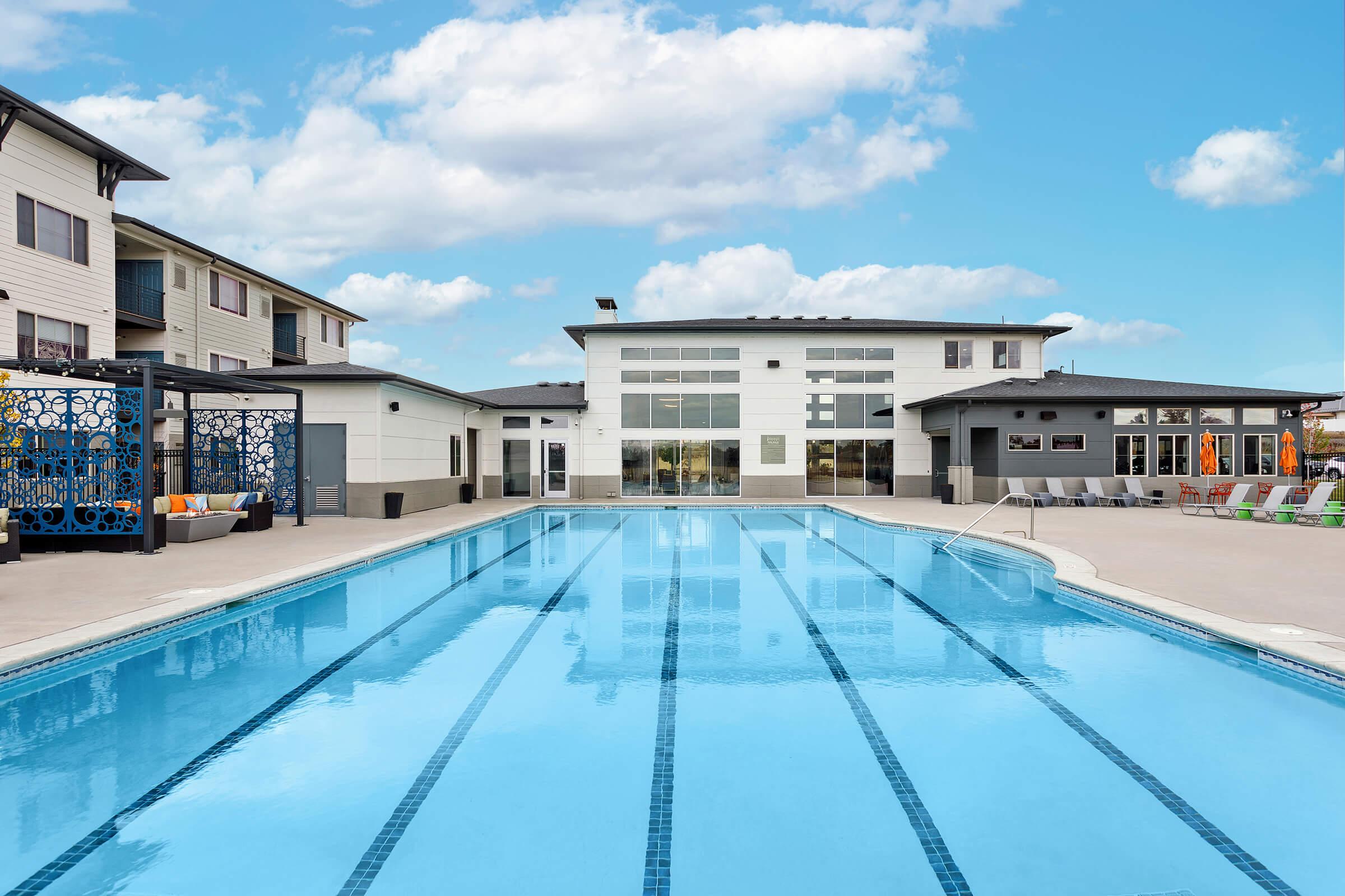 A clear blue swimming pool surrounded by a patio with lounge chairs, leading to a modern building with large windows. The sky is bright with clouds, and there are colorful cushions along the edge of the pool. The scene conveys a relaxing atmosphere in a residential community.