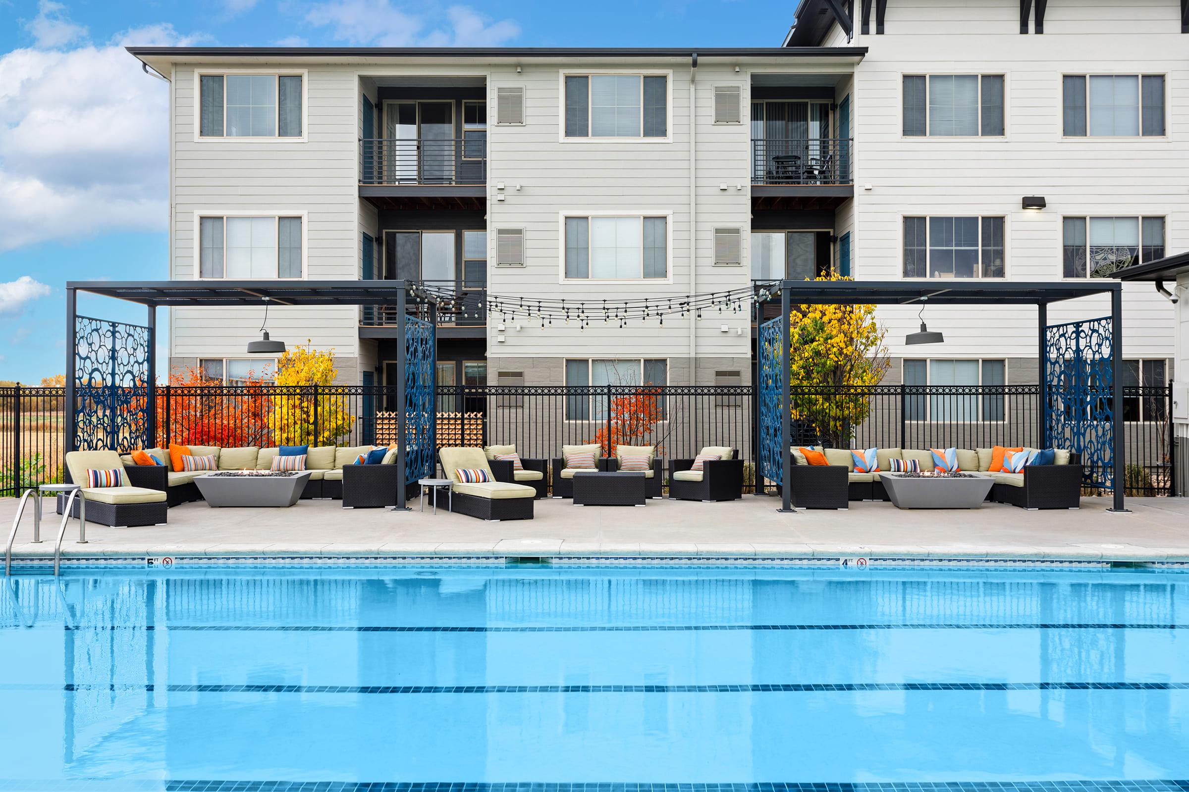 A modern apartment complex with a large outdoor pool. In the foreground, there are stylish seating areas with colorful cushions under shaded pergolas. The pool reflects the blue sky, and the background features several apartment buildings with fall foliage.