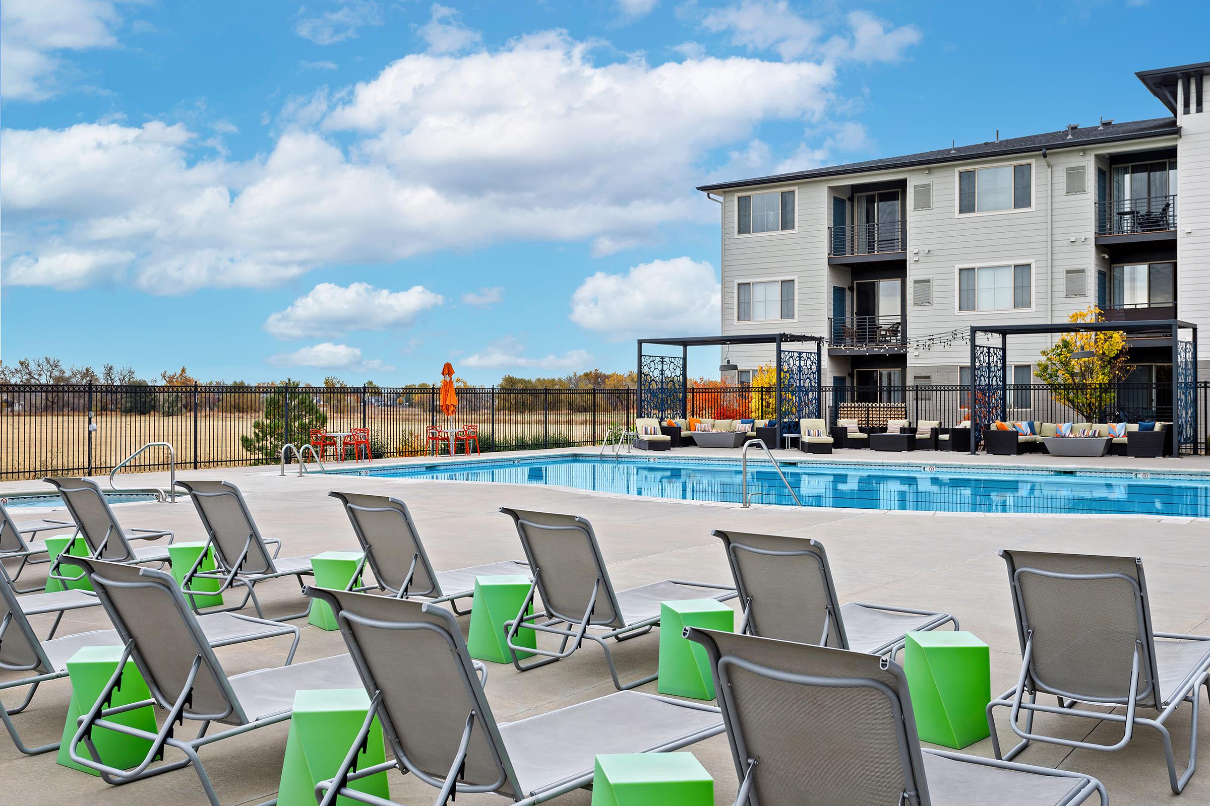 A modern outdoor swimming pool area surrounded by lounge chairs with green accents. In the background, an apartment building is visible, and the sky is bright with fluffy clouds. The pool area includes shaded cabanas and a landscaped space, creating a relaxing atmosphere for residents and guests.