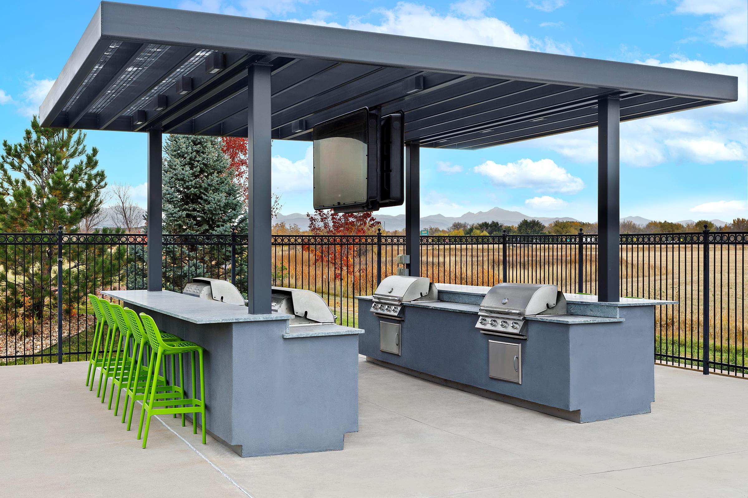 A modern outdoor grilling area featuring two stainless steel grills under a gray canopy. There's a long bar-style countertop with several bright green chairs and a scenic backdrop of trees and mountains in the distance, creating a vibrant outdoor setting.