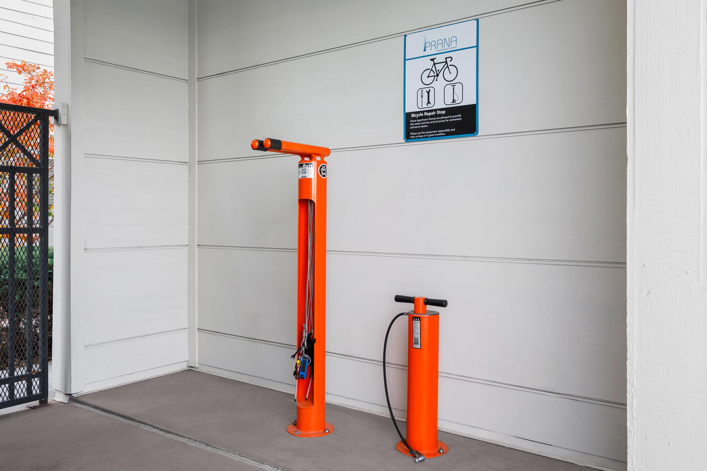 Bicycle repair station featuring an air pump and tools, set against a light-colored wall. A sign overhead provides instructions for using the repair tools. The station is brightly colored in orange, creating a vibrant contrast with the surrounding area.