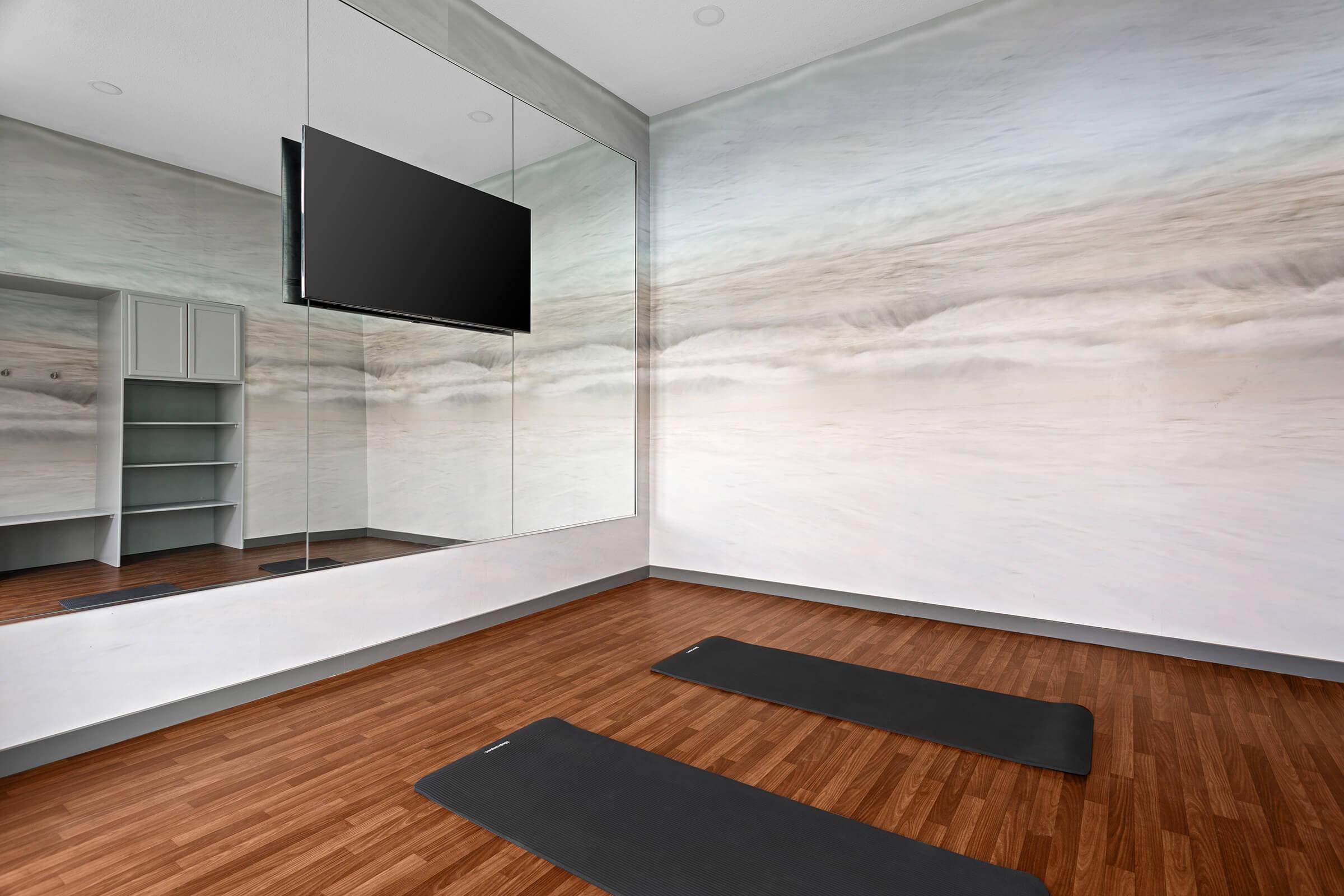 A serene yoga studio featuring two exercise mats on a wooden floor, large mirrors reflecting a calm ocean-themed mural, and a flat-screen TV mounted on the wall, creating a peaceful and inviting atmosphere for exercise or meditation.