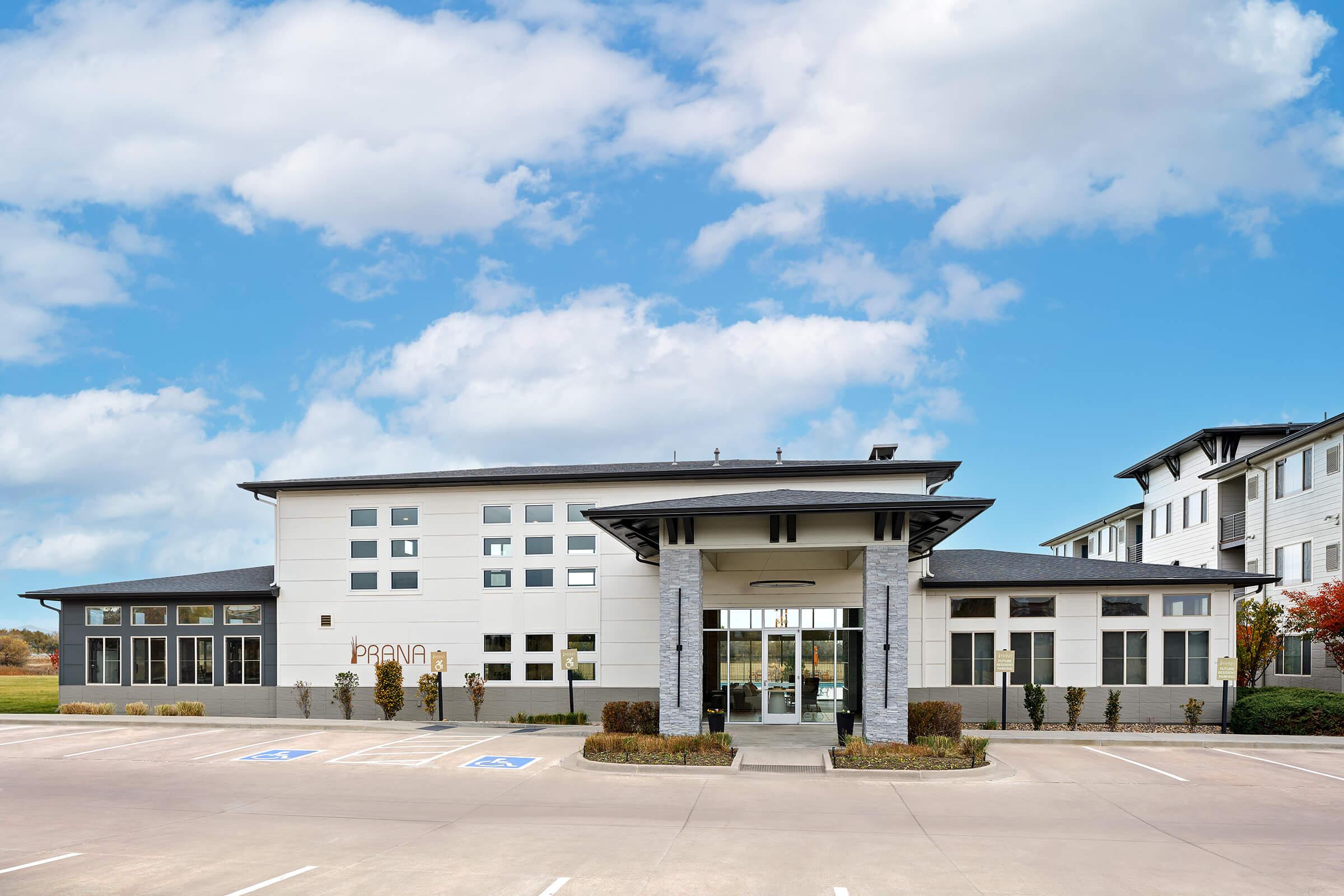 Modern building with a sleek design featuring large windows and a gray exterior, surrounded by landscaped areas. The entrance is framed by stone columns, and there are parking spaces in front. A clear blue sky with fluffy clouds is visible above, adding to the aesthetic appeal.