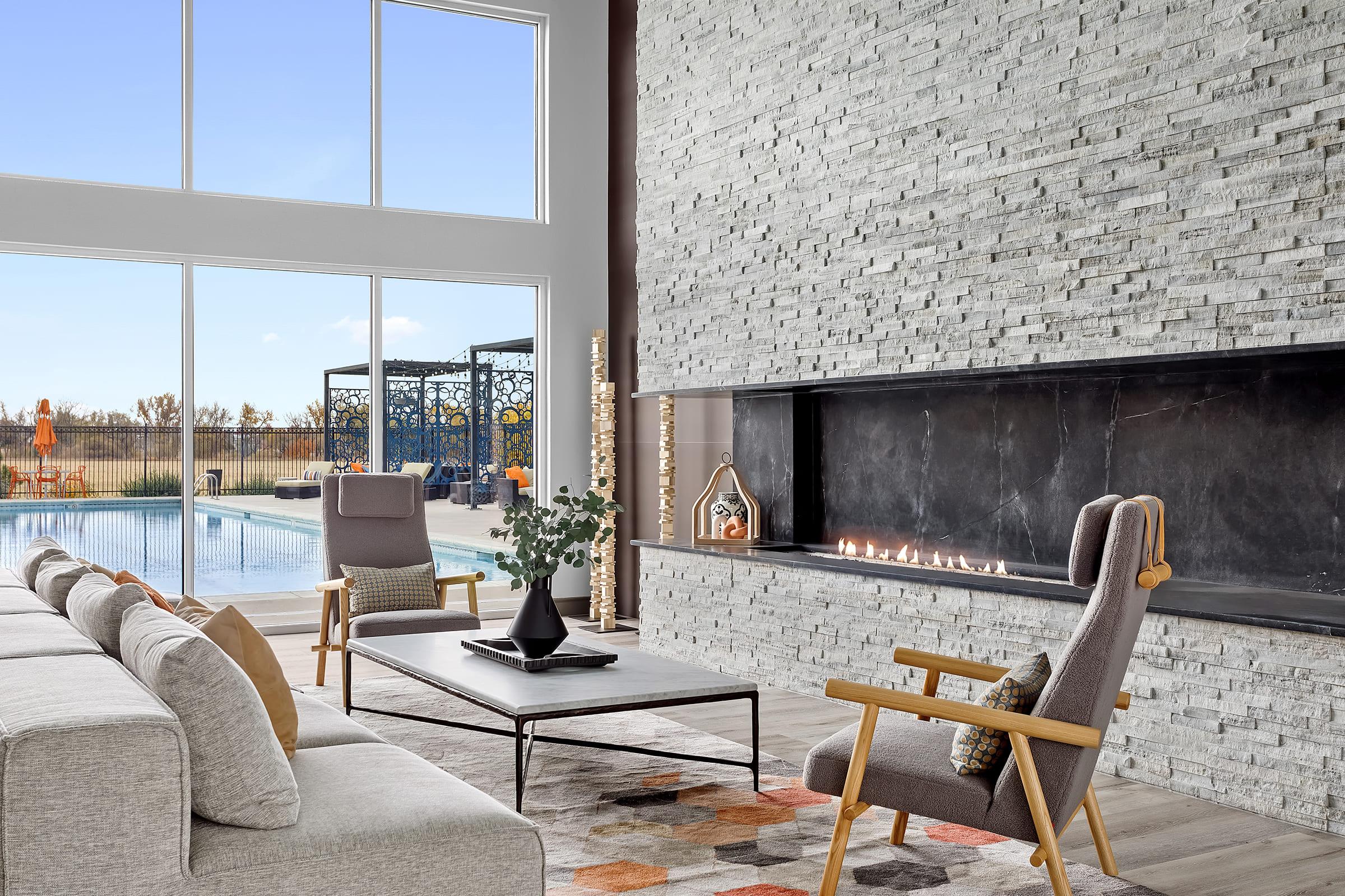 A modern living room featuring a large stone fireplace and a minimalist design. A comfortable sofa, two armchairs, and a coffee table are arranged around a colorful rug. Large windows provide a view of an outdoor pool area, enhancing the bright and airy atmosphere.
