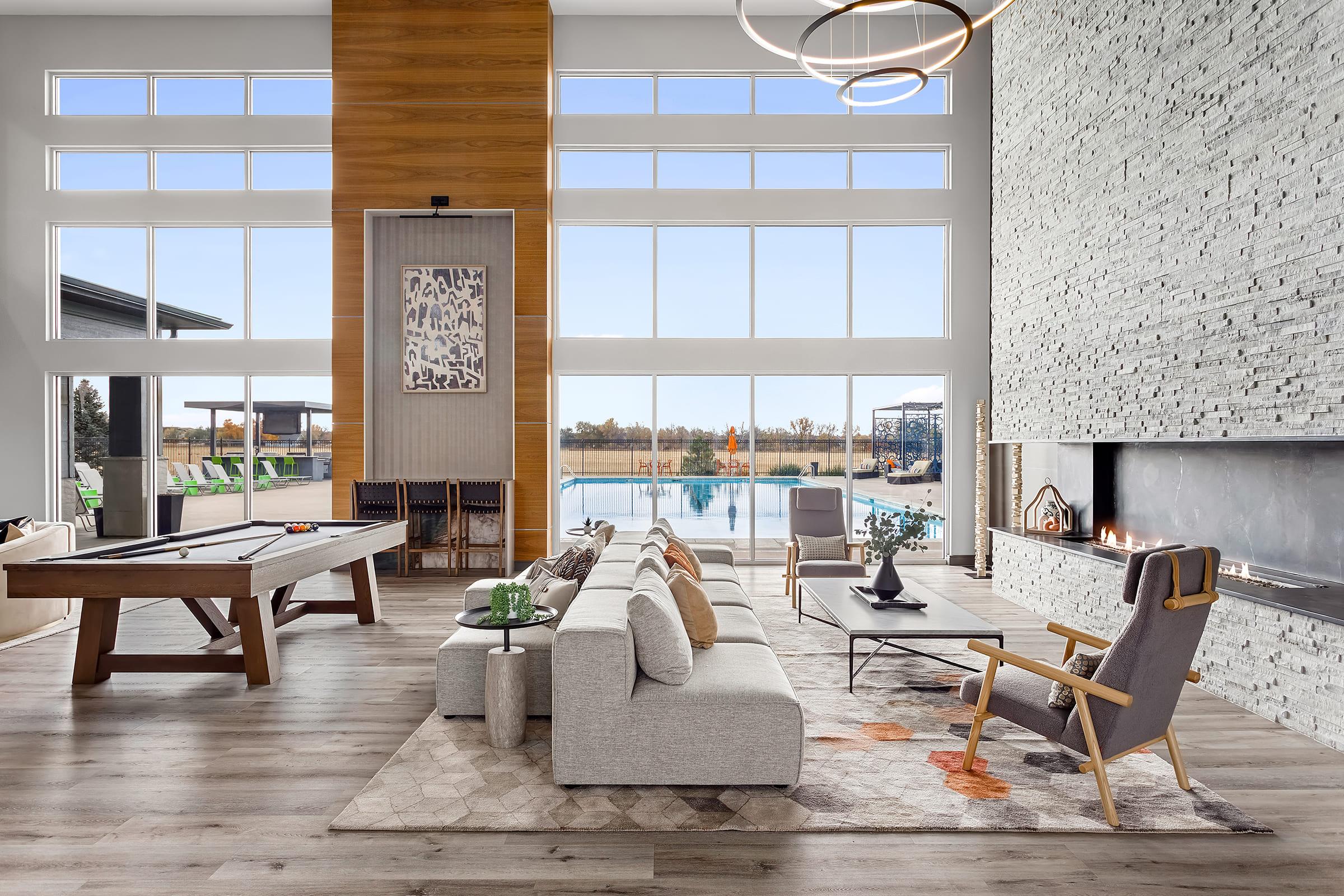 Modern living space featuring large windows with views of an outdoor pool. A stylish sectional sofa and armchair are arranged around a coffee table. A wooden pool table is positioned to one side, and a stone accent wall with a fireplace adds warmth to the room. Bright and airy atmosphere with neutral tones.