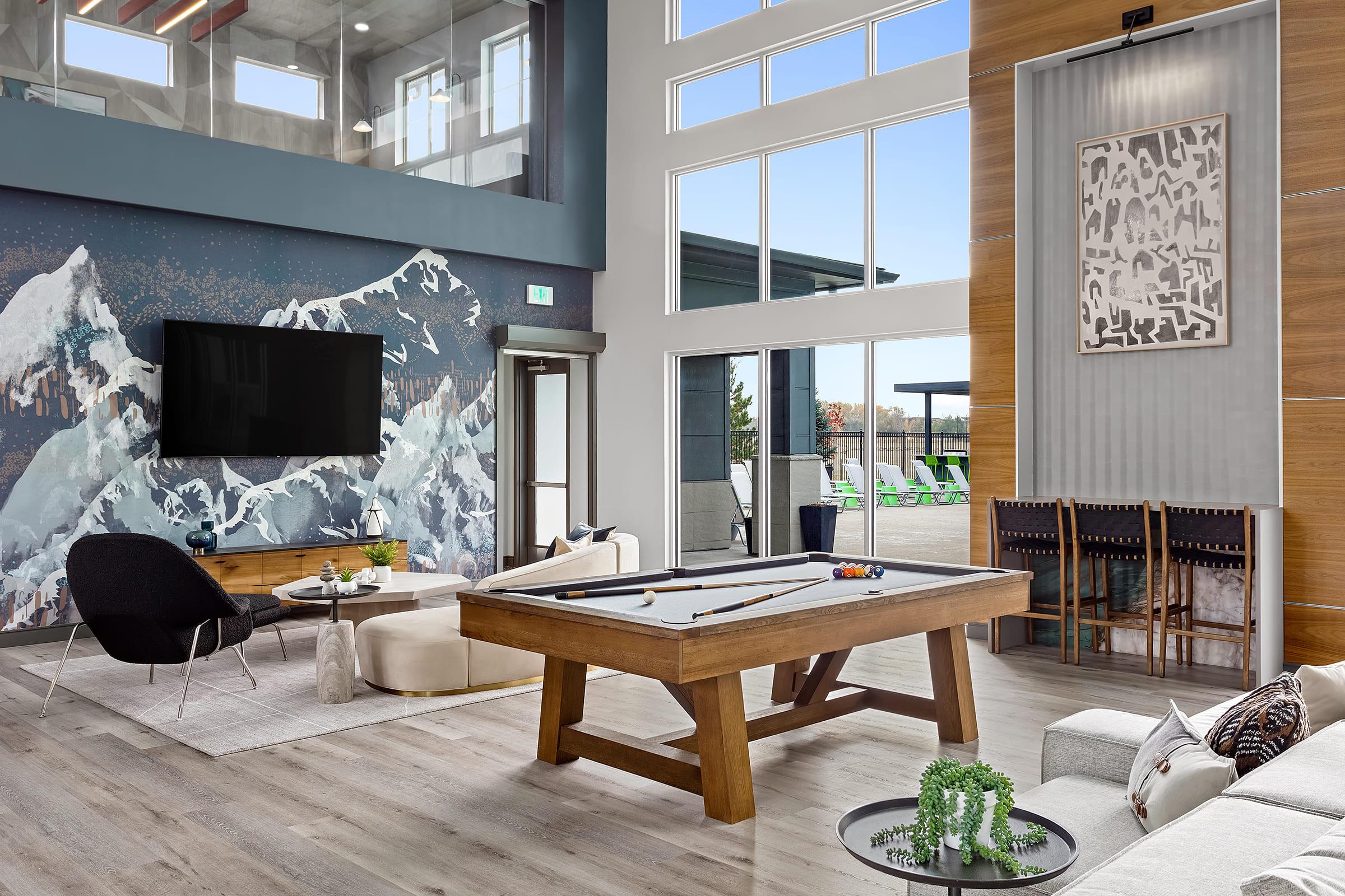 Interior view of a modern lounge area featuring a pool table, comfortable seating, and a large flat-screen TV. The wall is adorned with a mountain mural, and large windows allow natural light to fill the space. Stylish decor includes indoor plants and a contemporary art piece.