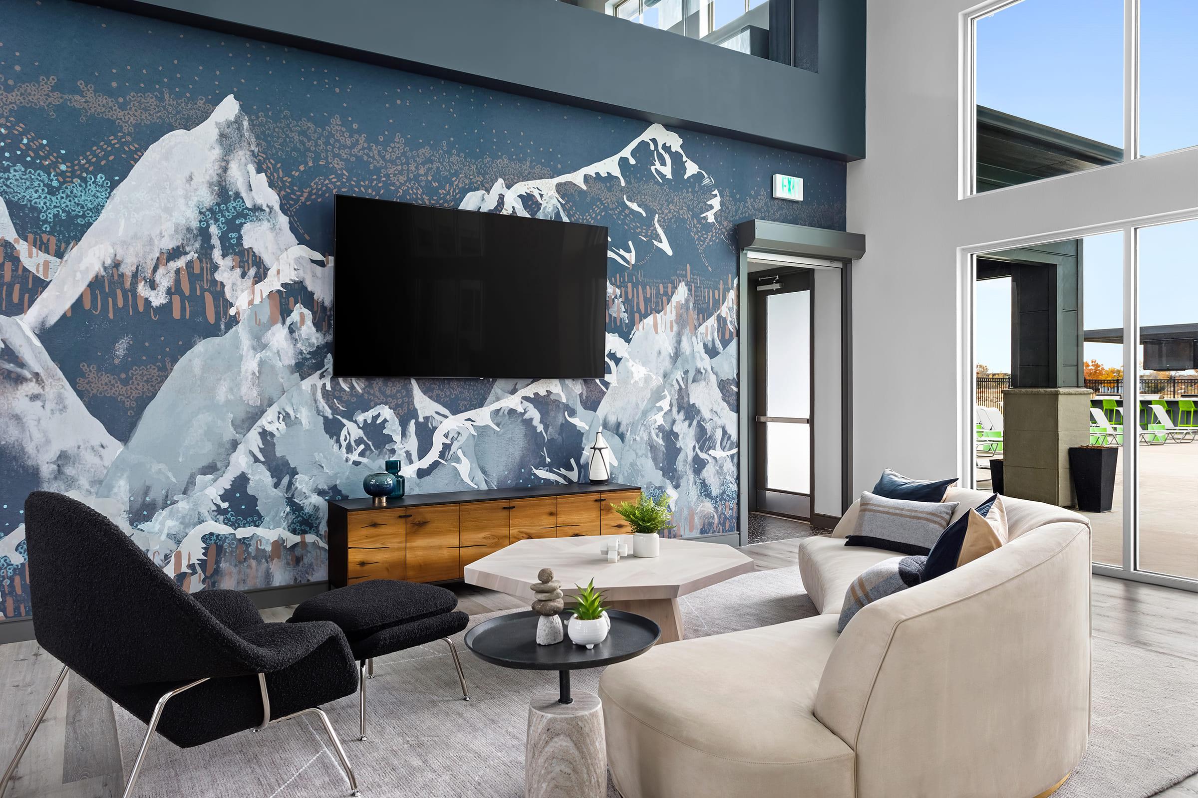 Modern living room featuring a large wall mural of snowy mountains. A sleek black television is mounted on the wall above a wooden console. The room includes stylish furniture: a black chair, a beige sofa, and a white coffee table, with large windows allowing natural light and offering a view of the outdoors.