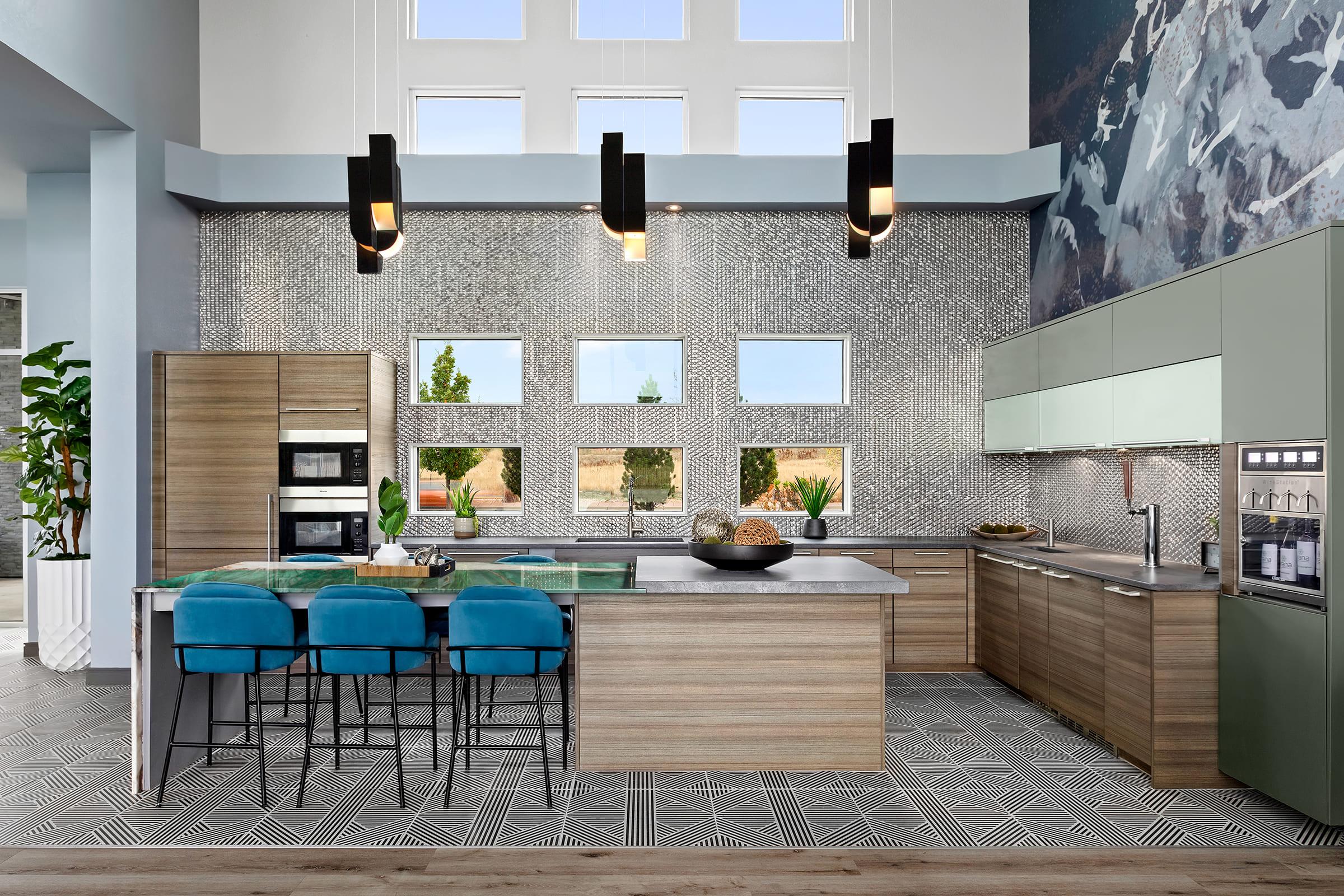 A modern kitchen featuring a large island with teal bar stools, sleek cabinetry in light wood and muted green, and contemporary appliances. The walls are adorned with unique tile patterns and large windows that provide ample natural light. Decorative plants add a touch of greenery.