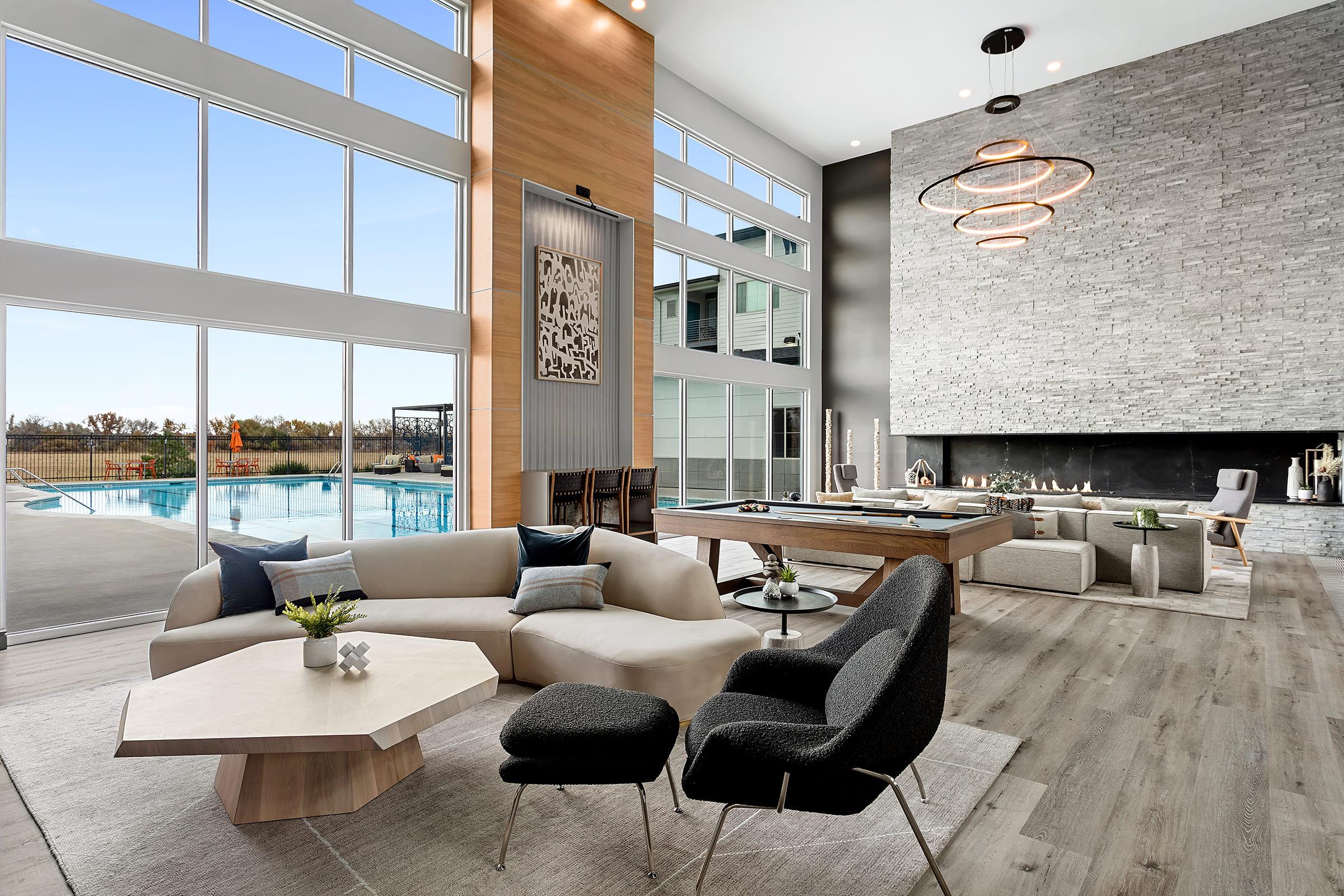 Modern living room with large windows showcasing an outdoor pool area. Features a light-colored sectional sofa and stylish black chairs around a geometric coffee table. A stone fireplace and minimalist decor enhance the contemporary vibe, complemented by a warm wood accent wall and elegant lighting fixtures.