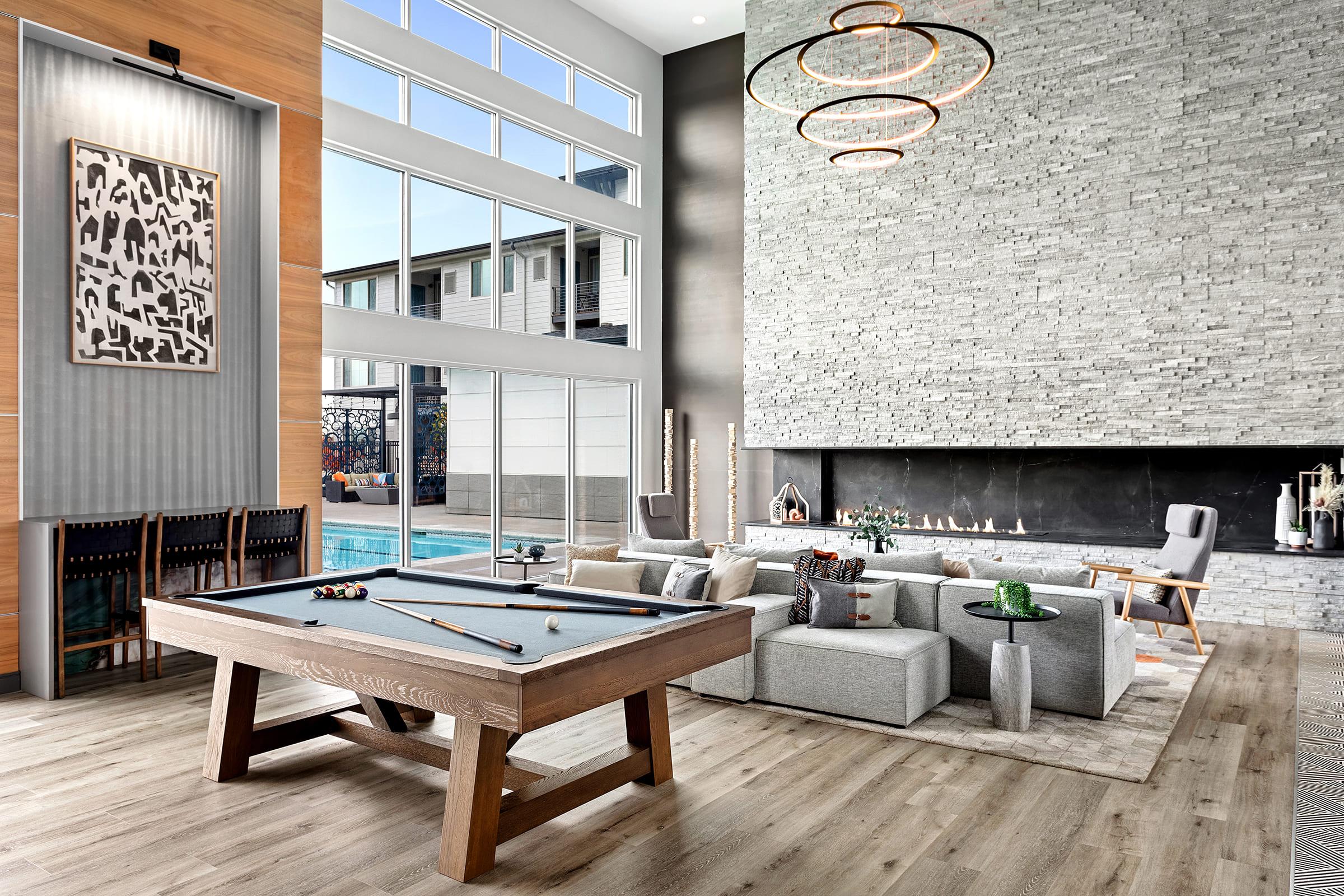 A luxurious modern living space featuring a sleek pool table, a cozy seating area with light gray sofas, and a contemporary fireplace. Large windows allow natural light to fill the room, while stylish decorations and a minimalist design create an inviting atmosphere. A swimming pool is visible outside.