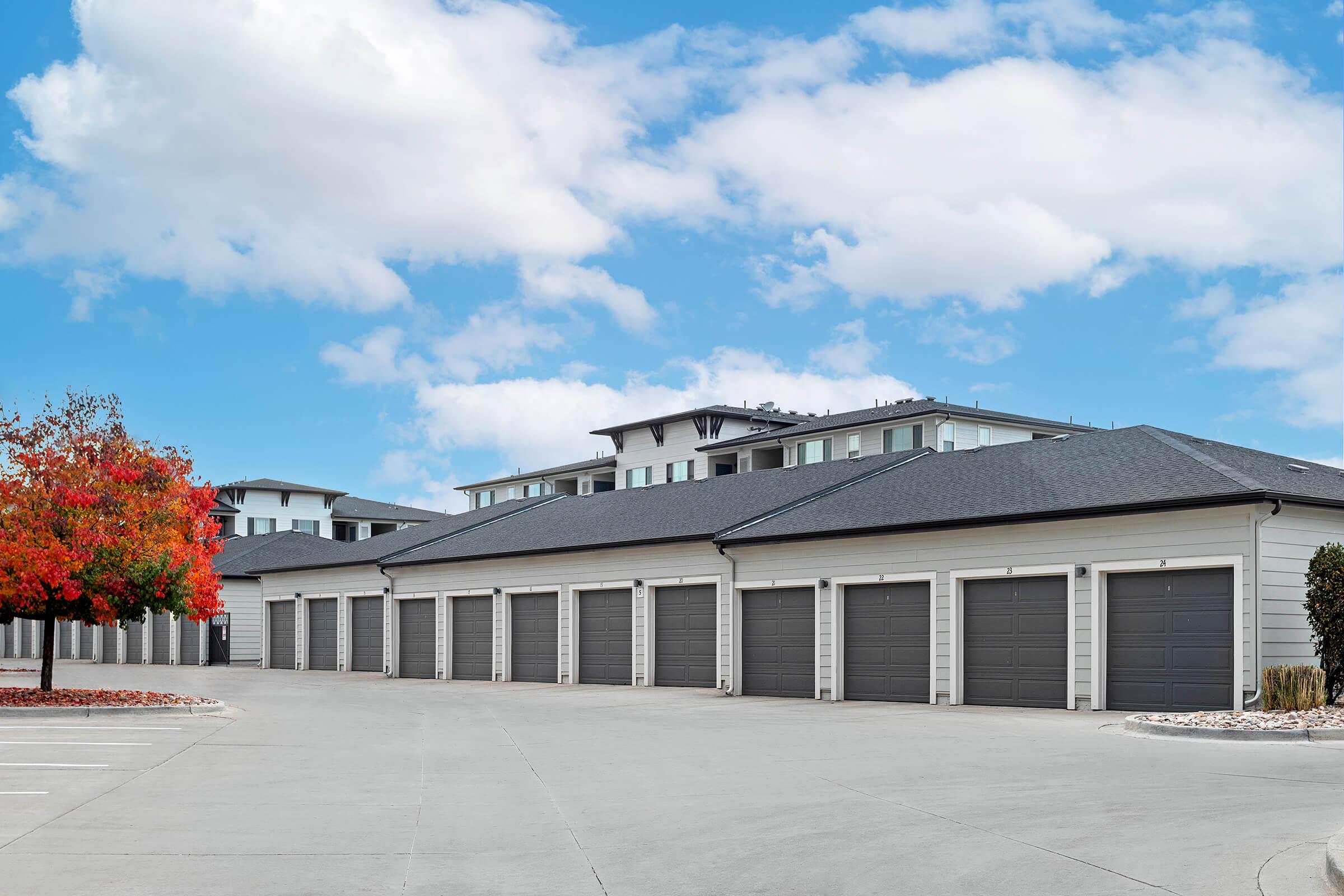 A row of gray garage doors lined up in a clean, paved area, with an apartment building visible in the background. A vibrant red tree adds a splash of color, contrasting with the gray surroundings. The sky is partly cloudy, creating a bright and inviting atmosphere.