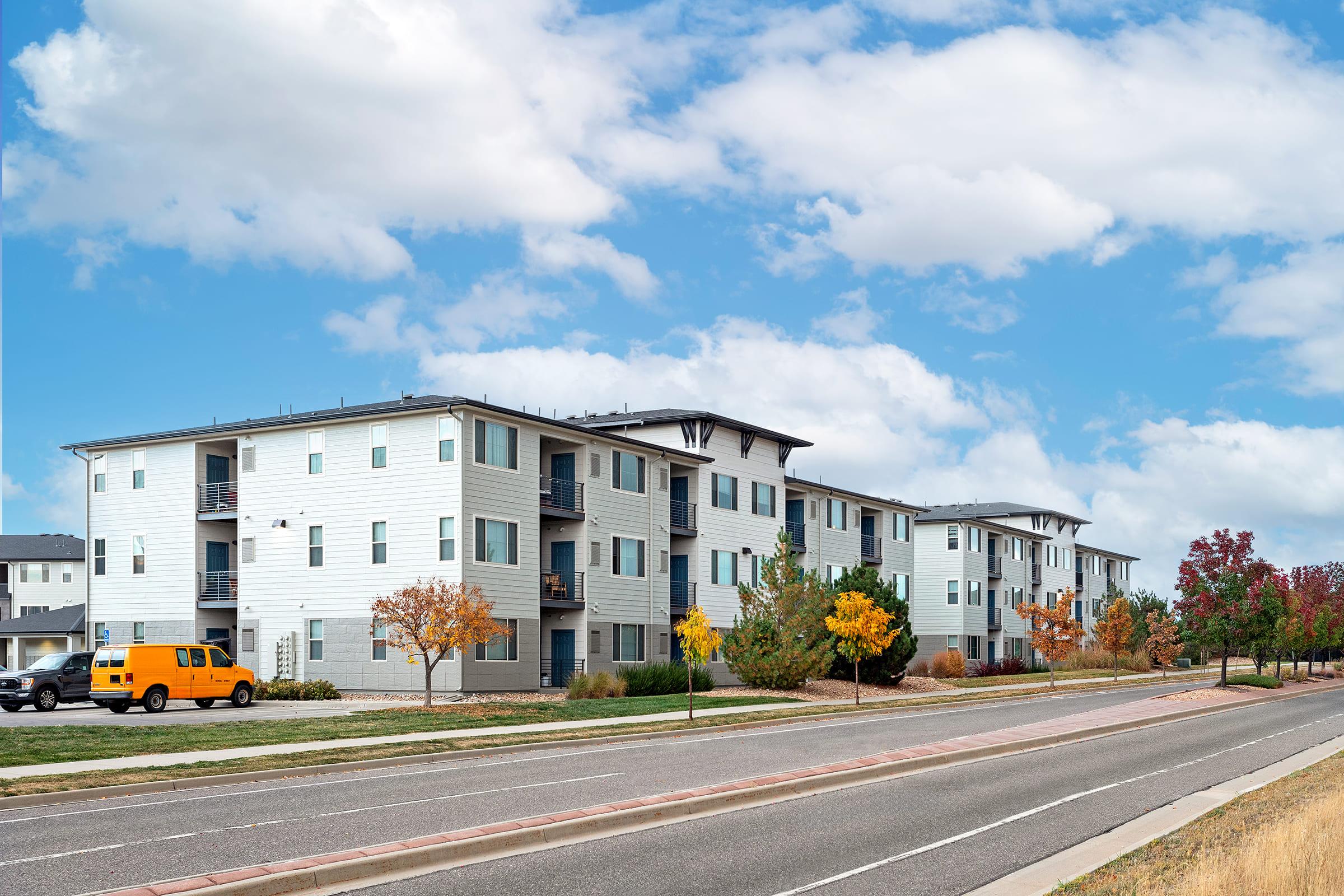 A row of modern apartment buildings with a light exterior, situated along a road. Fall foliage adorns the landscape with orange and yellow trees. A bright orange van is parked nearby, and the sky is partly cloudy with blue hues.