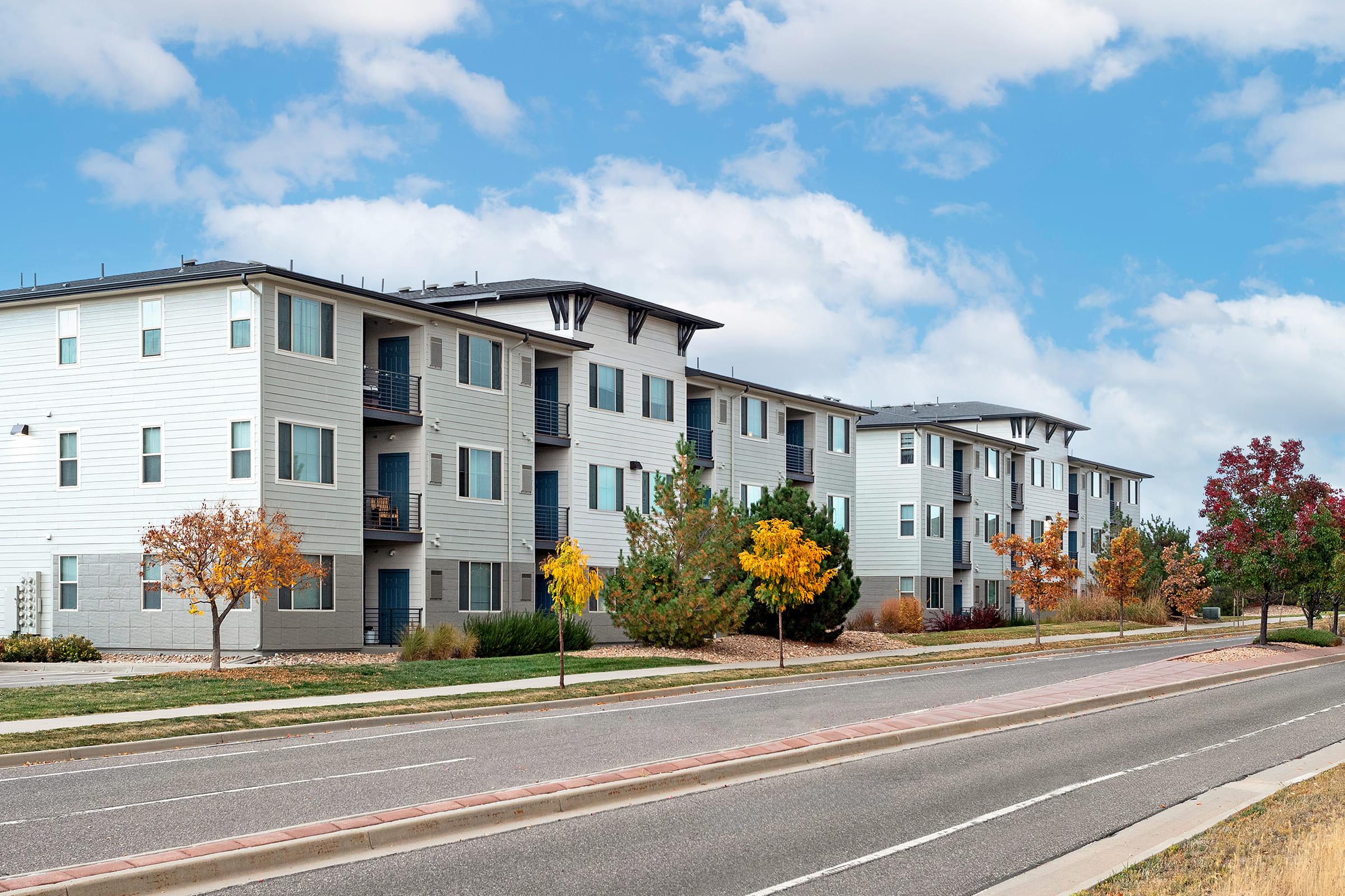 A row of modern apartment buildings with light-colored exteriors, featuring balconies and large windows. There are trees with autumn foliage in shades of yellow and red along a clean street, under a blue sky with scattered clouds. The scene captures a suburban neighborhood vibe.