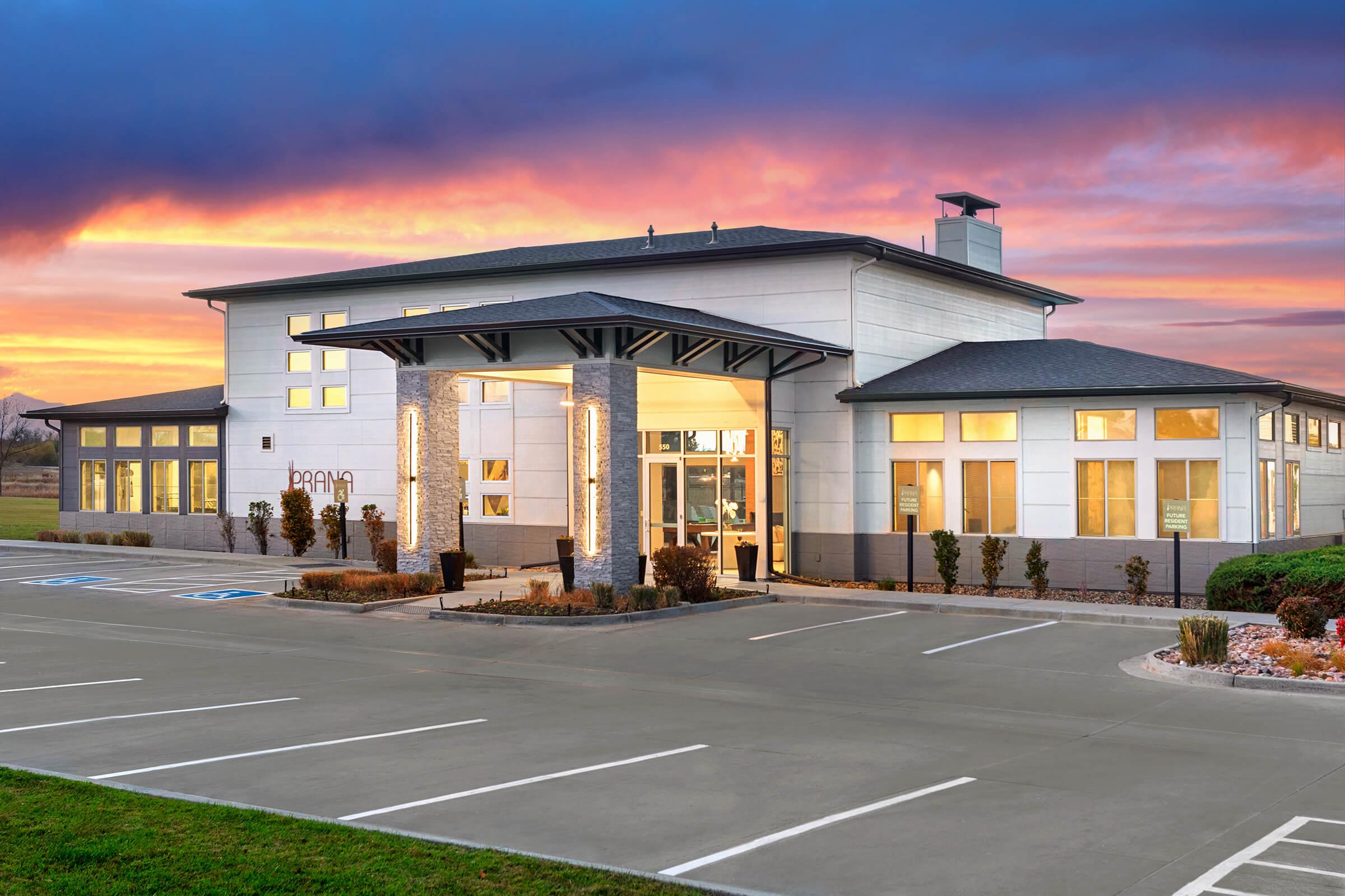 A modern building with a stylish facade, featuring large windows and a welcoming entrance. The structure is surrounded by a well-maintained parking lot and landscaping, with a colorful sunset sky in the background, creating a warm and inviting atmosphere.