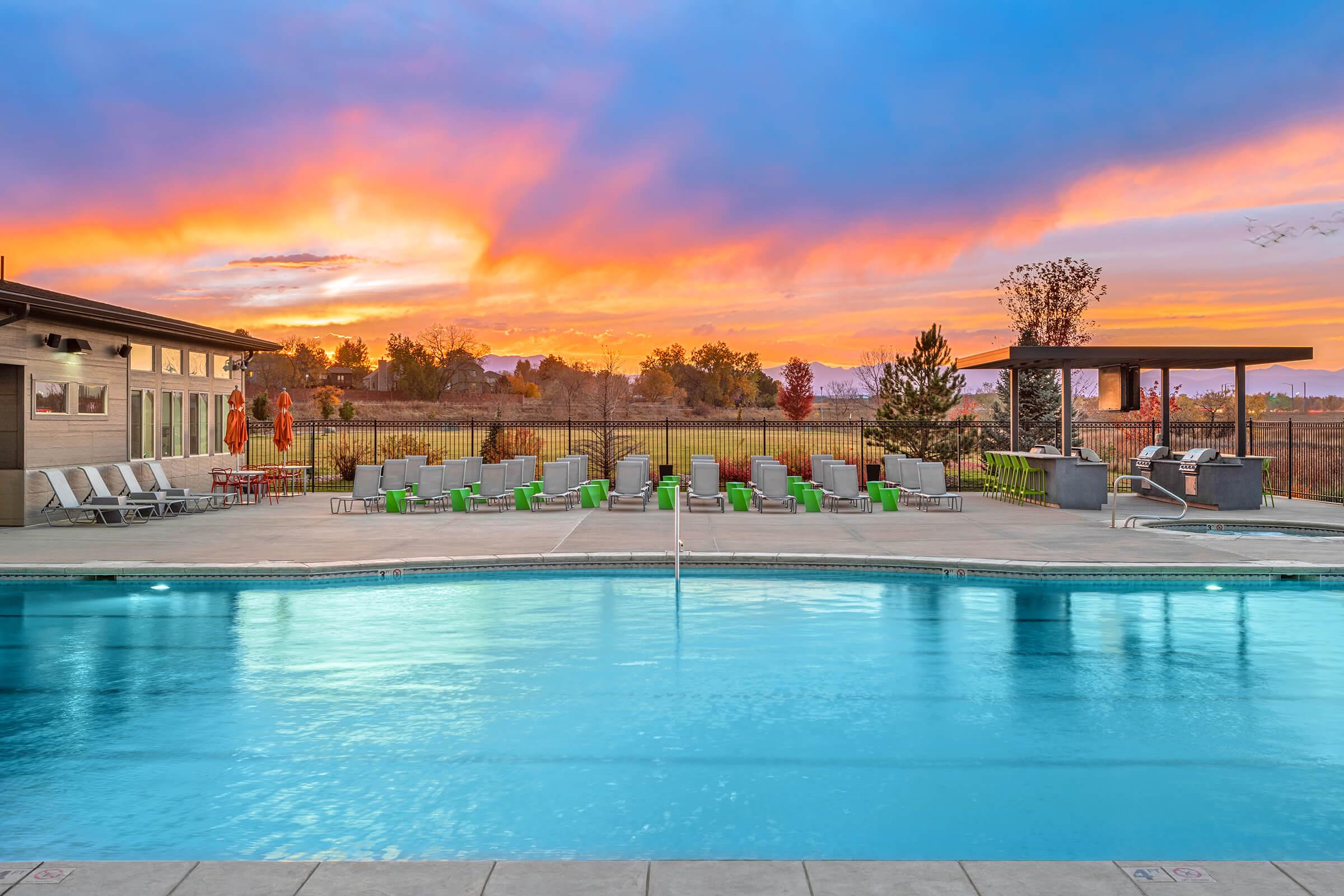 A tranquil outdoor pool area at sunset, featuring clear blue water and a backdrop of vibrant orange and purple skies. Surrounding the pool are green lounge chairs and a cozy seating area, with a pergola and landscaped greenery in the distance, creating a relaxing atmosphere.