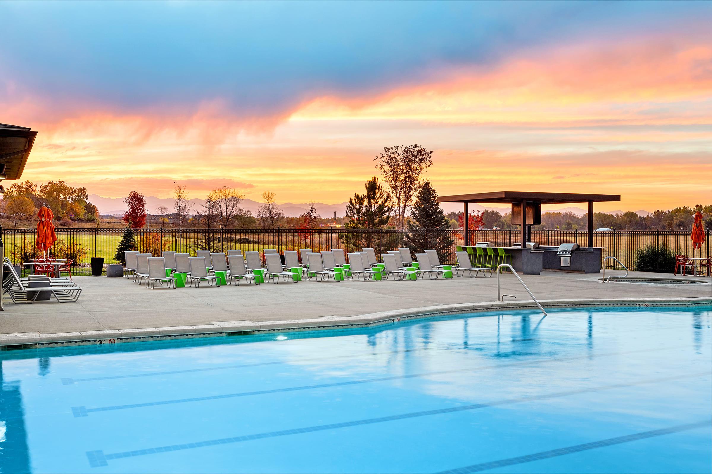 A serene outdoor swimming pool surrounded by lounge chairs and a shaded area with a barbecue grill. The sunset sky features vibrant colors, and the landscape includes trees and mountains in the distance, creating a tranquil atmosphere for relaxation and enjoyment.
