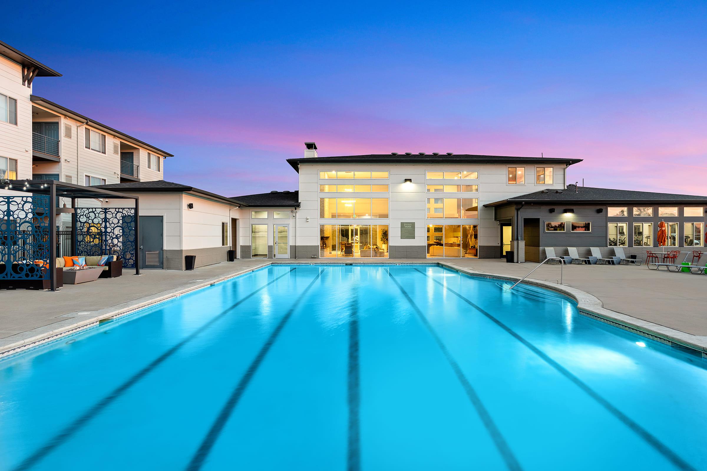 A modern pool area at twilight, featuring a clear blue swimming pool with reflections of the surrounding buildings. Stylish lounge chairs and patio furniture are visible by the poolside, alongside a contemporary building with large windows glowing softly in the evening light.