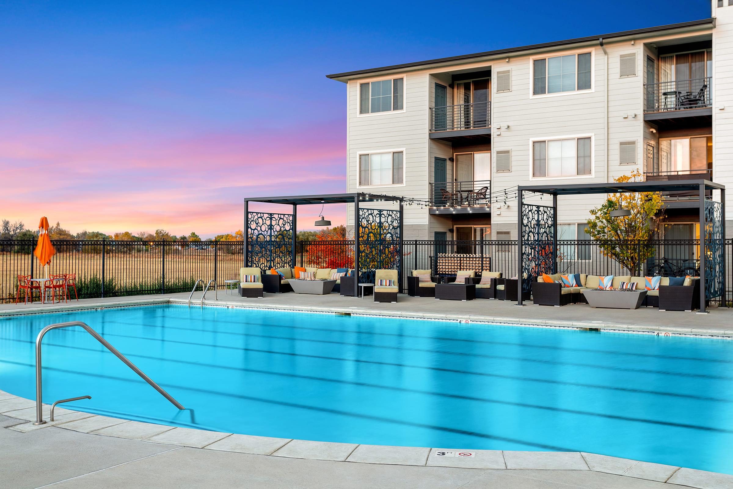 A sparkling blue swimming pool surrounded by lounge seating, shaded areas, and colorful decor. Modern apartment buildings are in the background, and a beautiful sunset sky adds a warm glow to the scene.