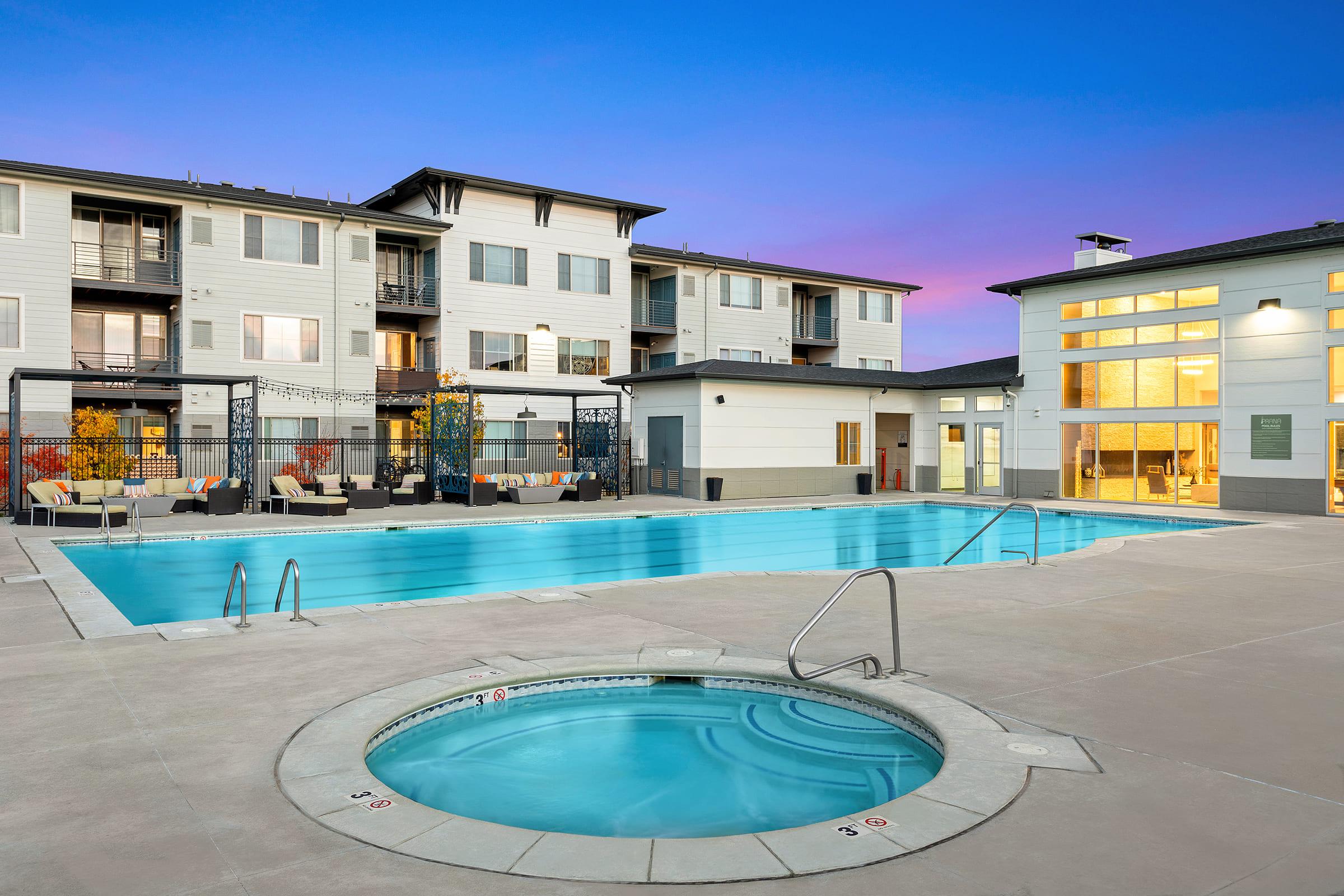 A modern apartment complex with a large swimming pool and a heated jacuzzi. The pool area is surrounded by lounge chairs and decorative landscaping, while the buildings feature balconies and large windows. The sky is a vibrant gradient of blue and purple, indicating sunset.