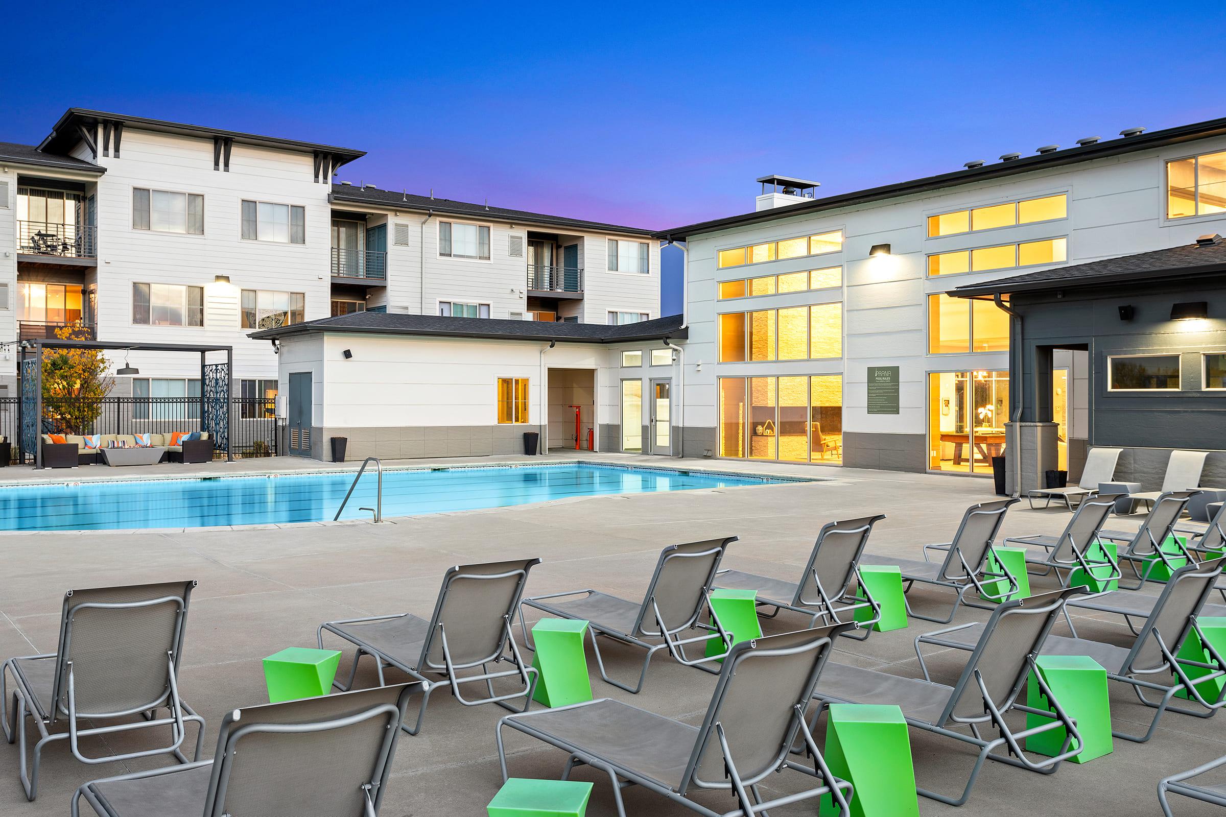 A modern apartment complex with a swimming pool surrounded by lounge chairs. The pool area is well-lit, and the buildings feature large windows that create a welcoming atmosphere. The sky is a vibrant blue, suggesting a twilight setting. Green accents on the chairs add a pop of color.