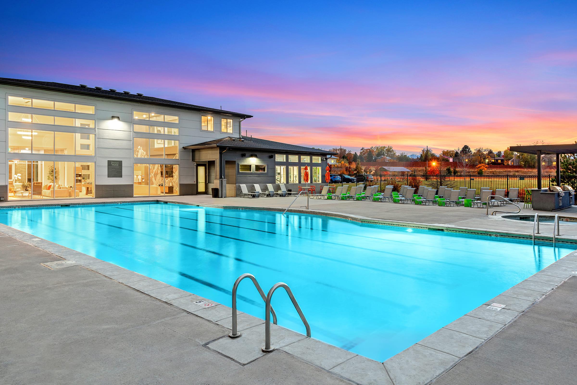 A clear blue swimming pool surrounded by lounge chairs, with a modern building featuring large windows in the background. The sky is painted in vibrant shades of purple and orange as the sun sets, creating a serene and inviting atmosphere.
