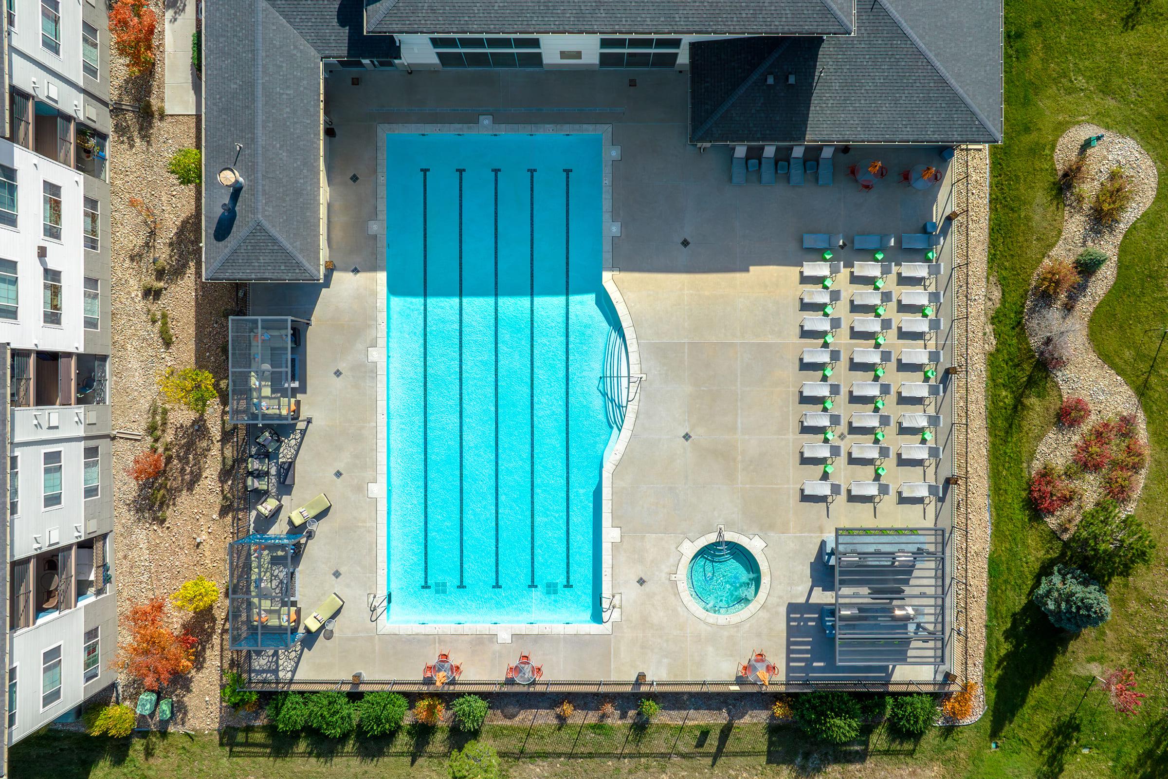 Aerial view of a swimming pool with clear blue water, featuring several lanes marked out. Surrounding the pool are lounge chairs and a hot tub. The area is landscaped with trees and decorative stones, and there are residential buildings nearby.