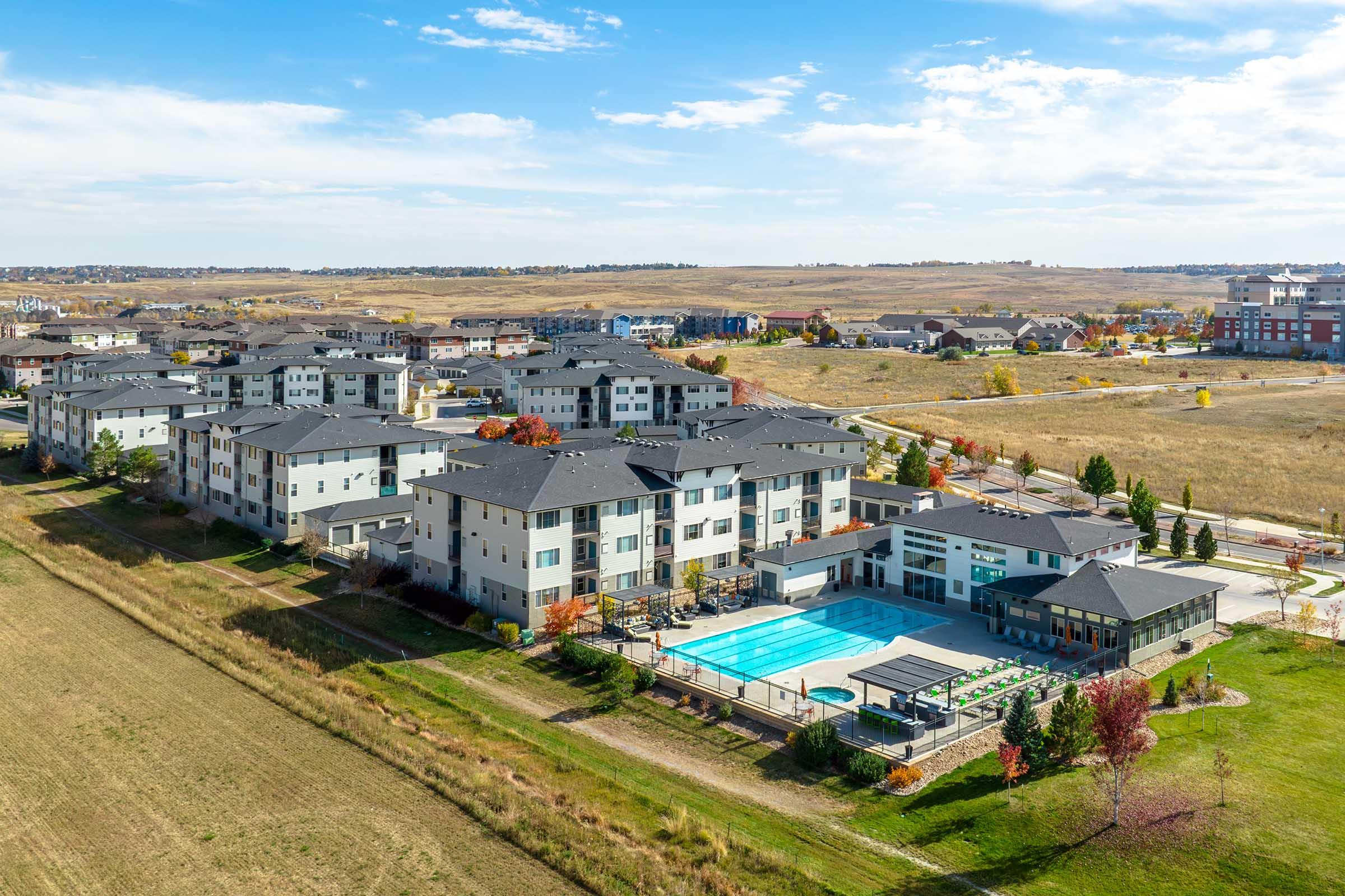 Aerial view of a modern residential complex featuring multiple buildings, a swimming pool, and landscaped areas. The surrounding landscape includes open fields and additional housing in the distance, with trees displaying autumn colors under a blue sky.