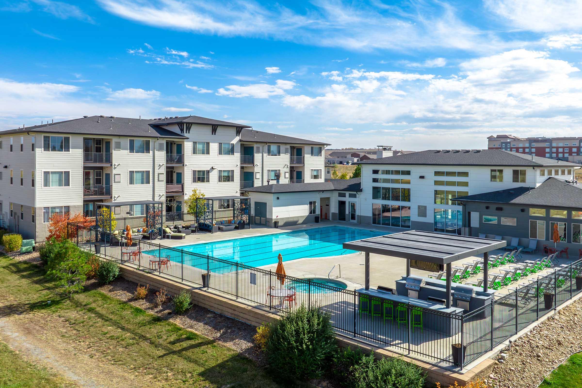 Aerial view of a modern apartment complex with a large swimming pool surrounded by lounge chairs and umbrellas. The buildings are three stories high, featuring balconies and greenery around the area. The sky is clear with some clouds, creating a bright atmosphere.