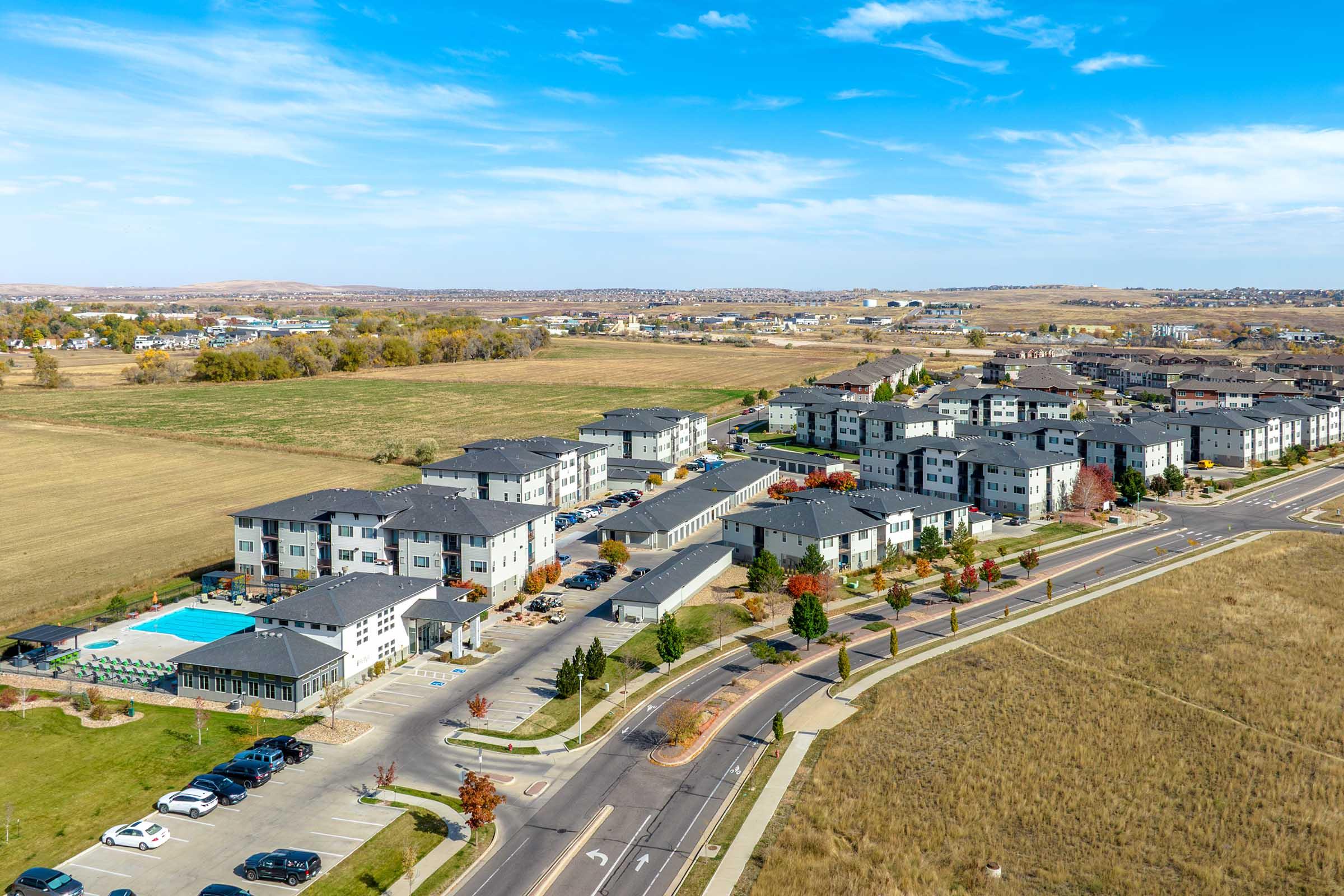 Aerial view of a modern apartment complex surrounded by green fields, featuring multiple buildings with flat roofs, landscaped areas, a swimming pool, and parking spaces along the streets. The background shows additional residential structures and open spaces under a clear blue sky.