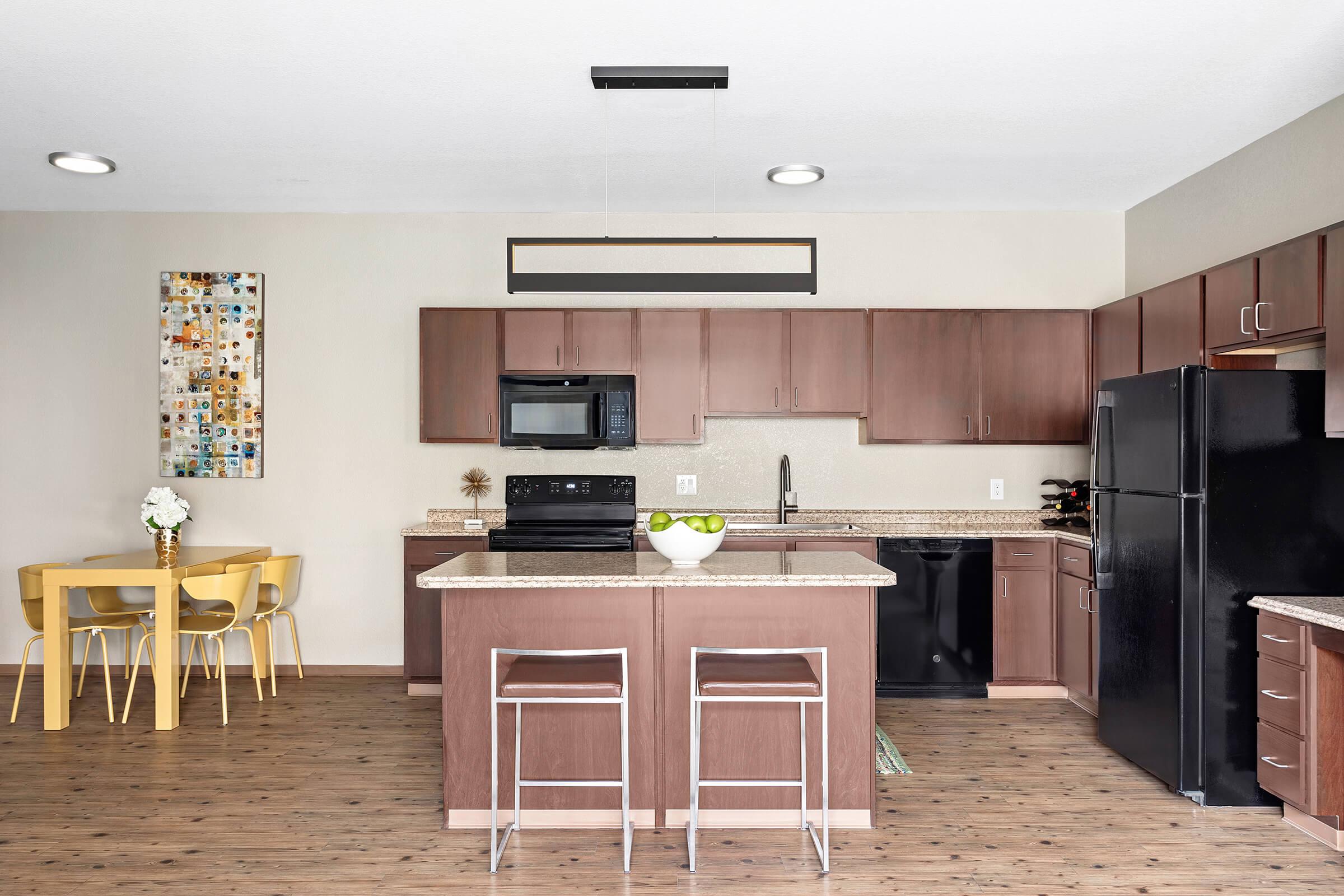 Modern kitchen featuring wooden cabinetry, granite countertops, and stainless steel appliances. An island with bar stools is in the center, alongside a dining area with a yellow table and chairs. A bowl of green apples sits on the island, and artwork is displayed on the wall.