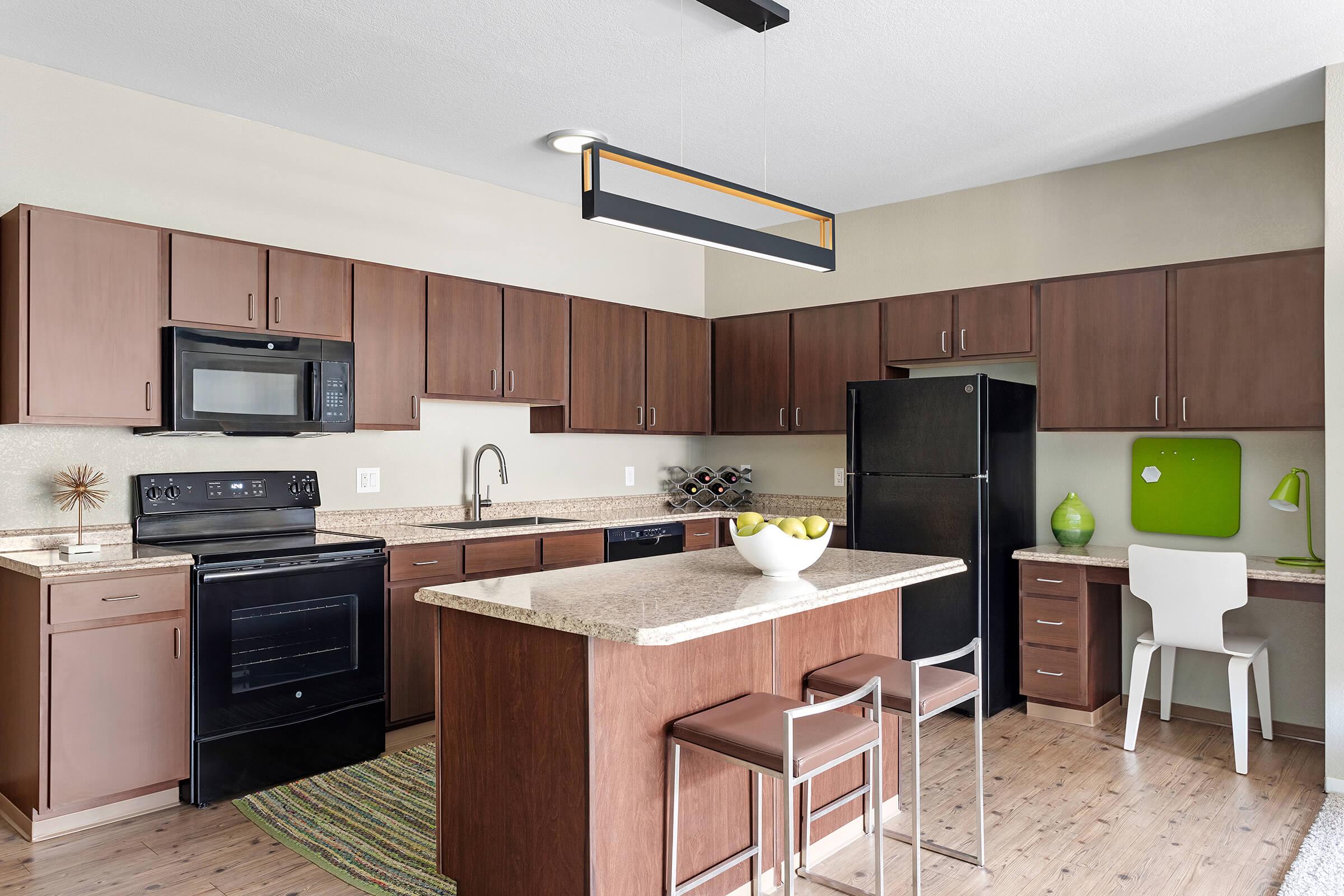 Modern kitchen with dark wood cabinetry and stainless steel appliances. Features an island with a granite countertop and seating, a microwave, and a black refrigerator. A bowl of green apples is on the island, and there’s a small desk area with a green accent wall. Bright and spacious atmosphere.